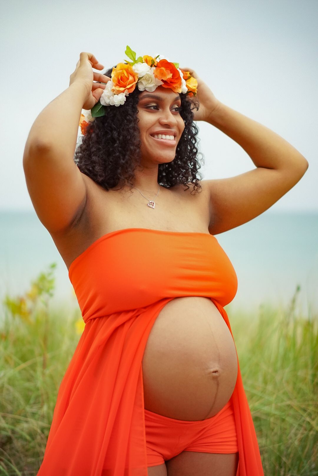 Maternity photo of pregnant woman in orange dress with floral crown against natural backdrop.