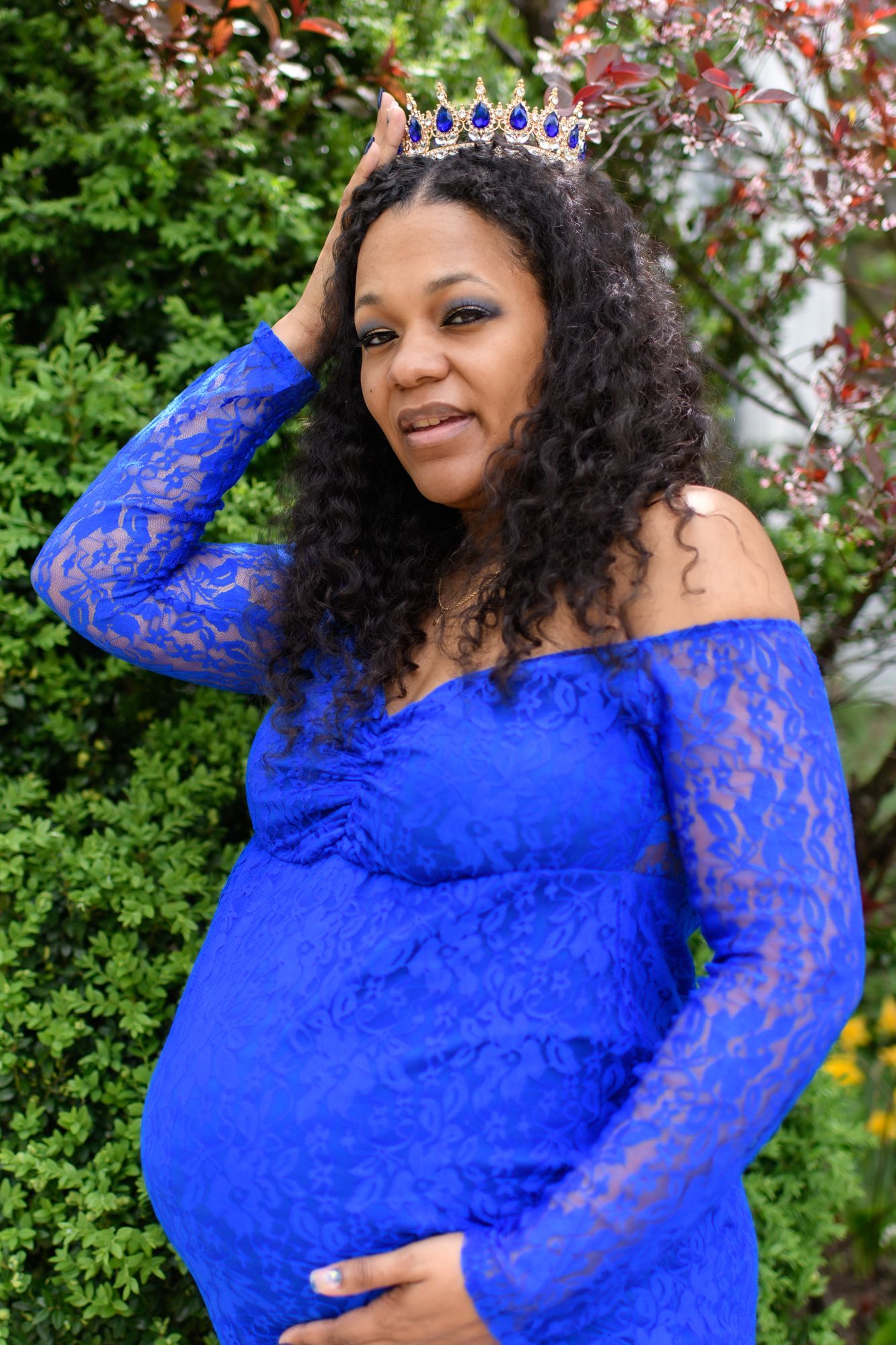 Maternity photoshoot in royal blue lace dress with crown against garden backdrop.