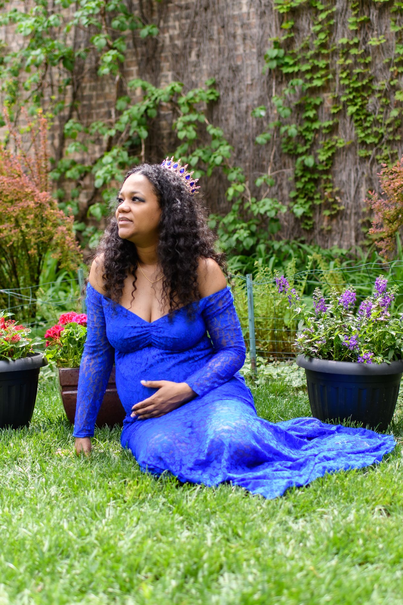 Maternity session showing woman in blue lace gown reclining on grass with flower garden.
