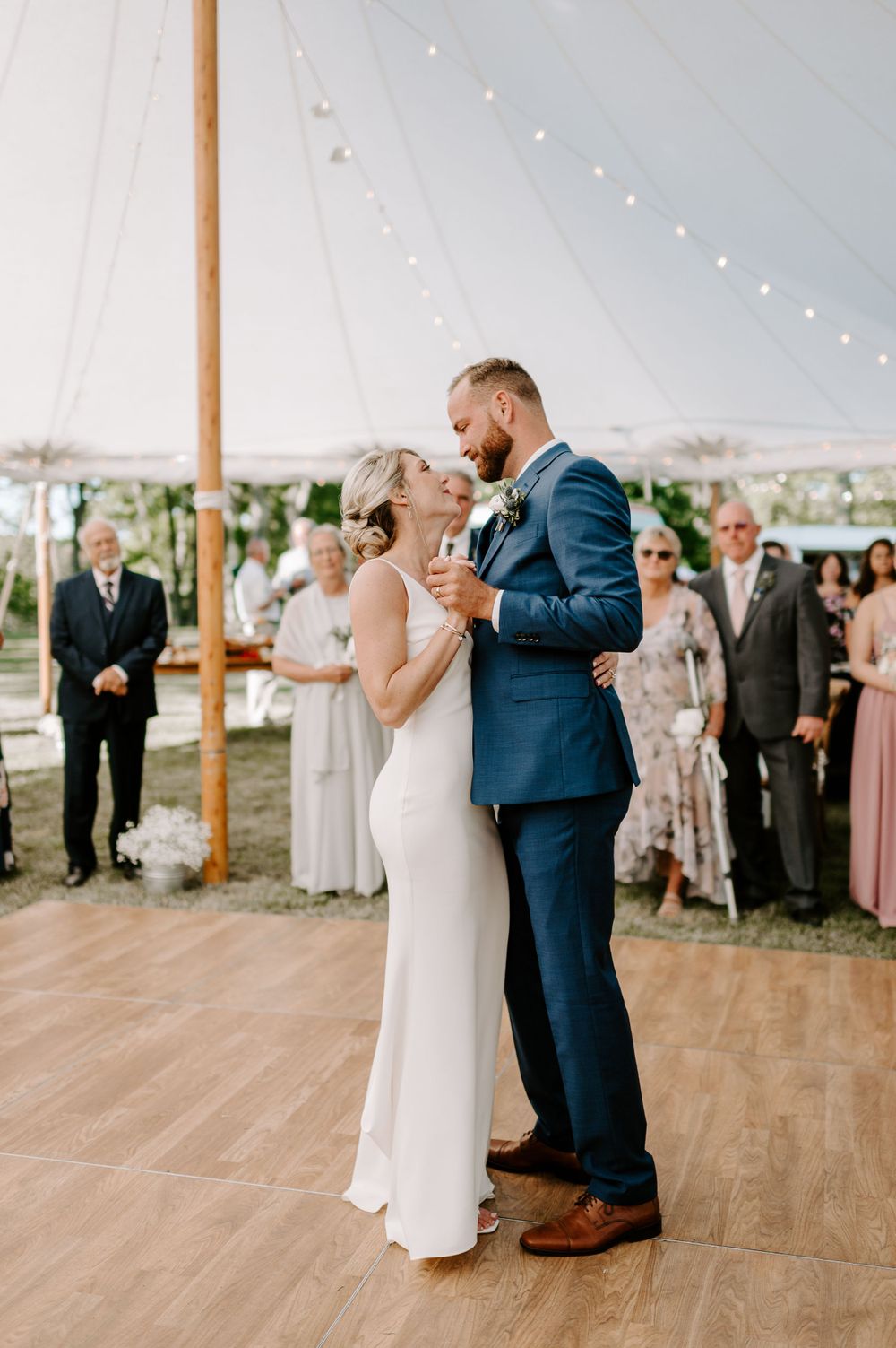 Newlyweds dancing together on wooden floor under white tent with string lights and guests watching.