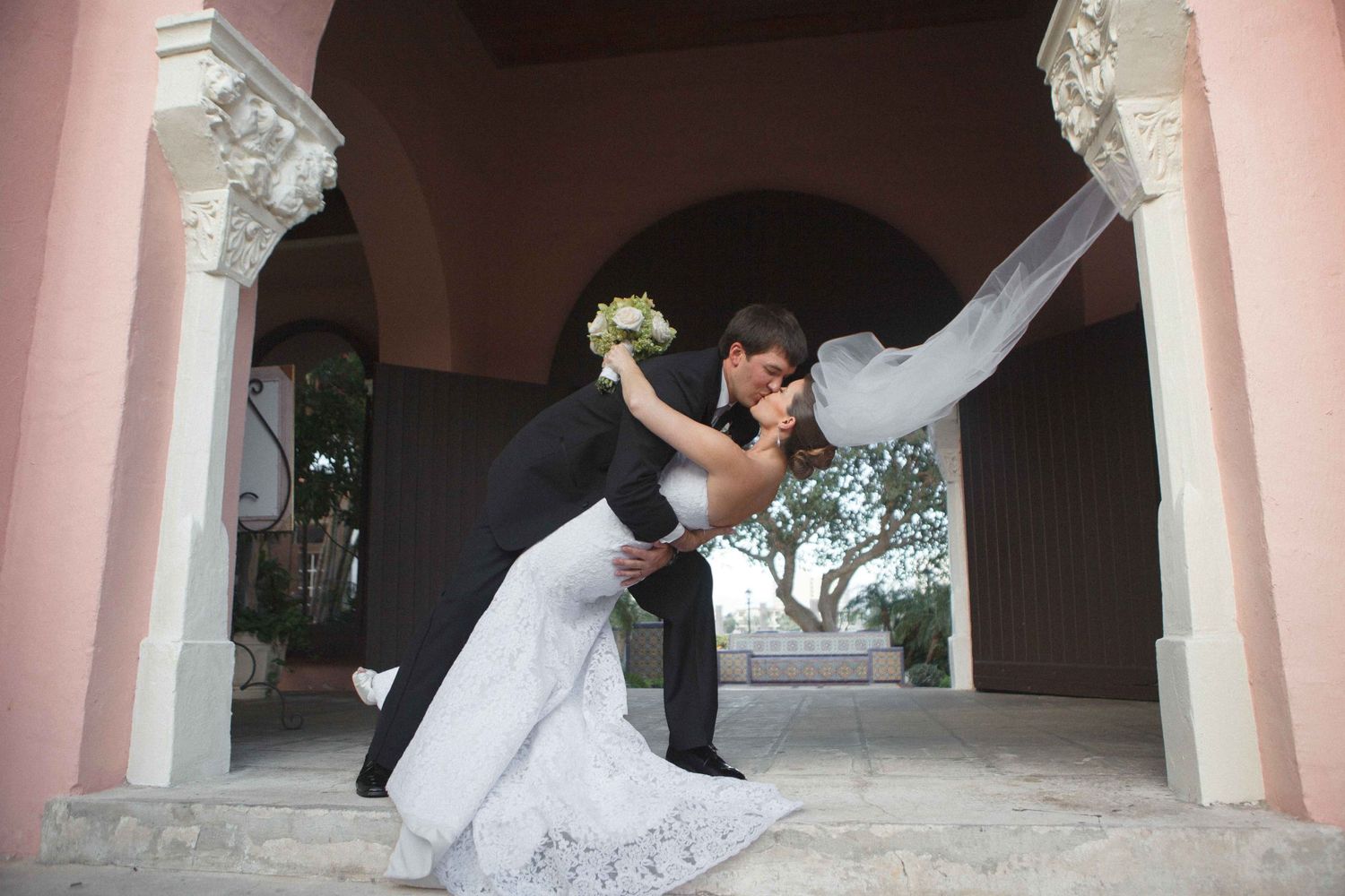 A bride and groom share a romantic kiss in front of pink columns at their wedding.