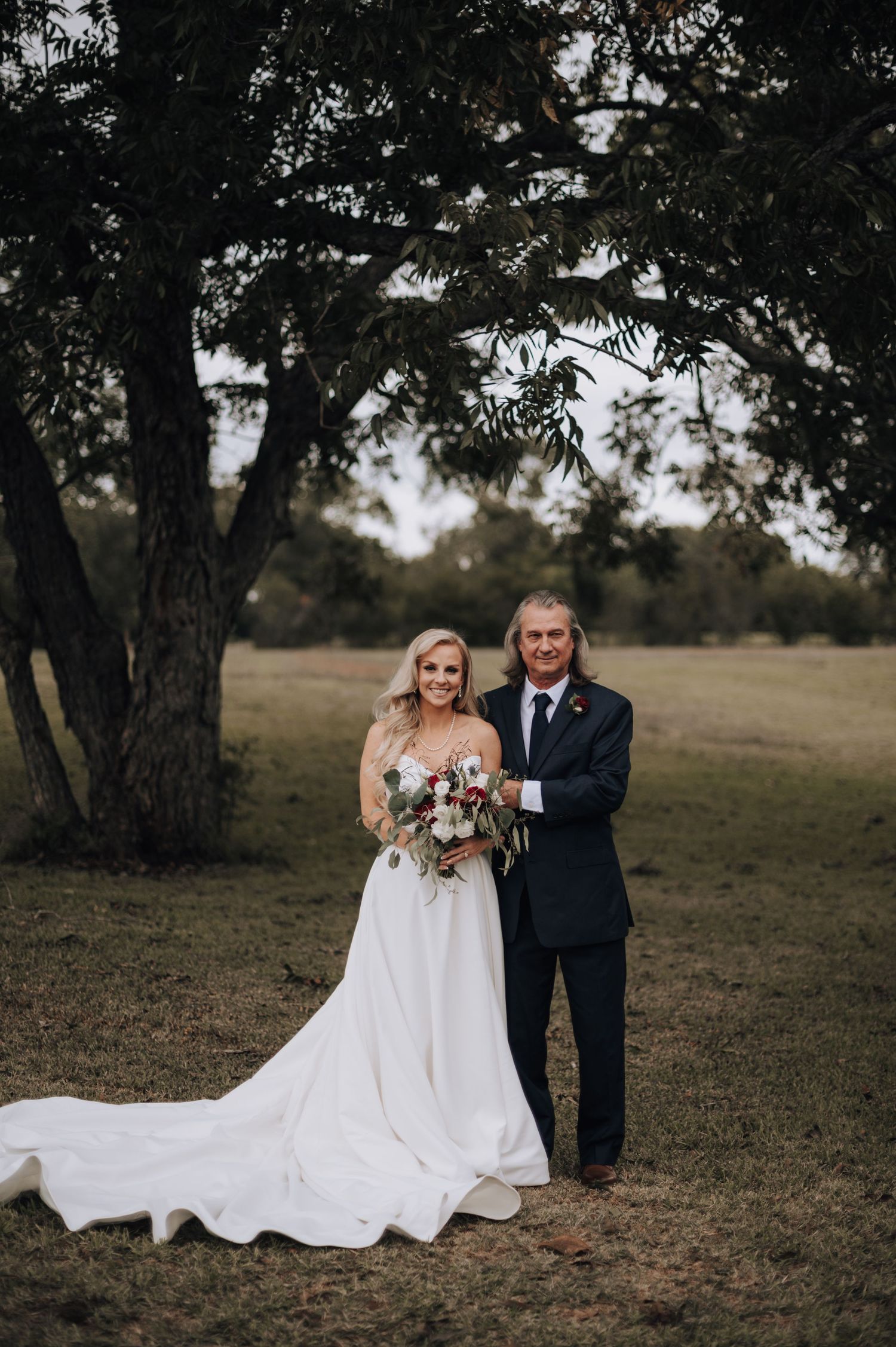 Wedding couple poses under large tree in rustic outdoor setting with flowing white gown.