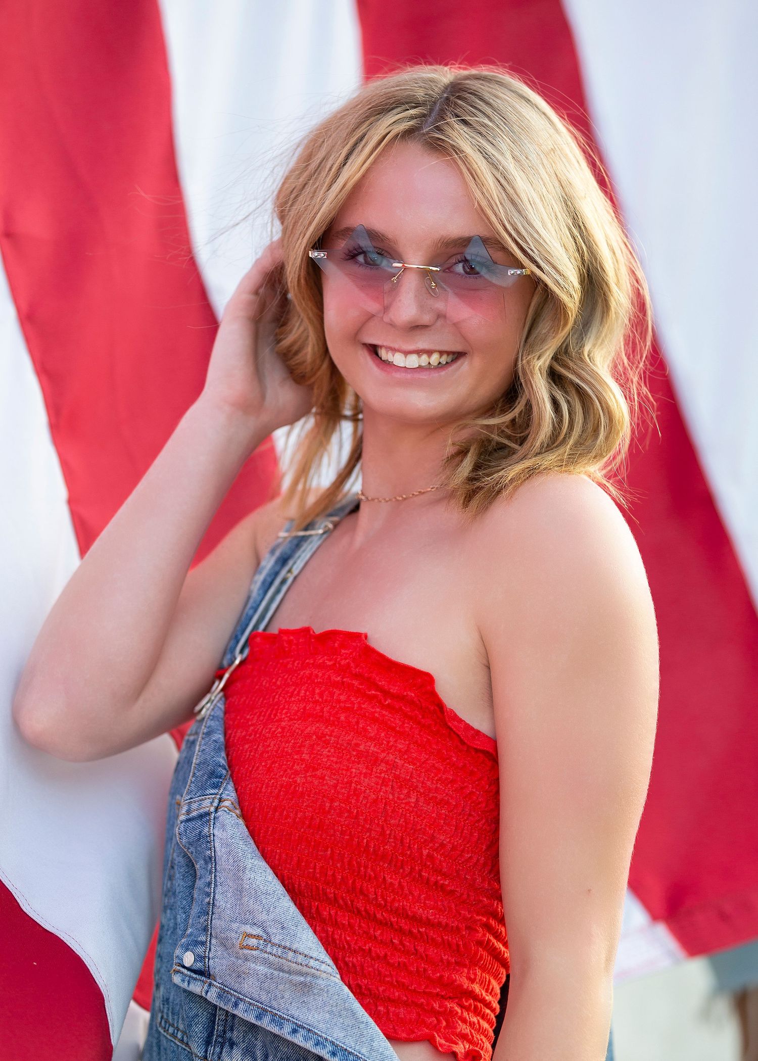 close shot of senior girl posed in front of the American flag smiling with star sunglasses on and touching hair