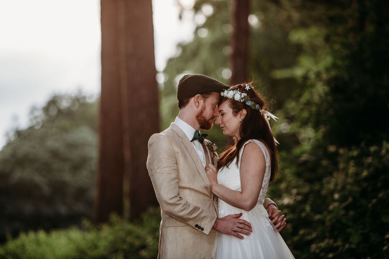 A tender moment between newlyweds beneath tall trees in soft natural lighting.