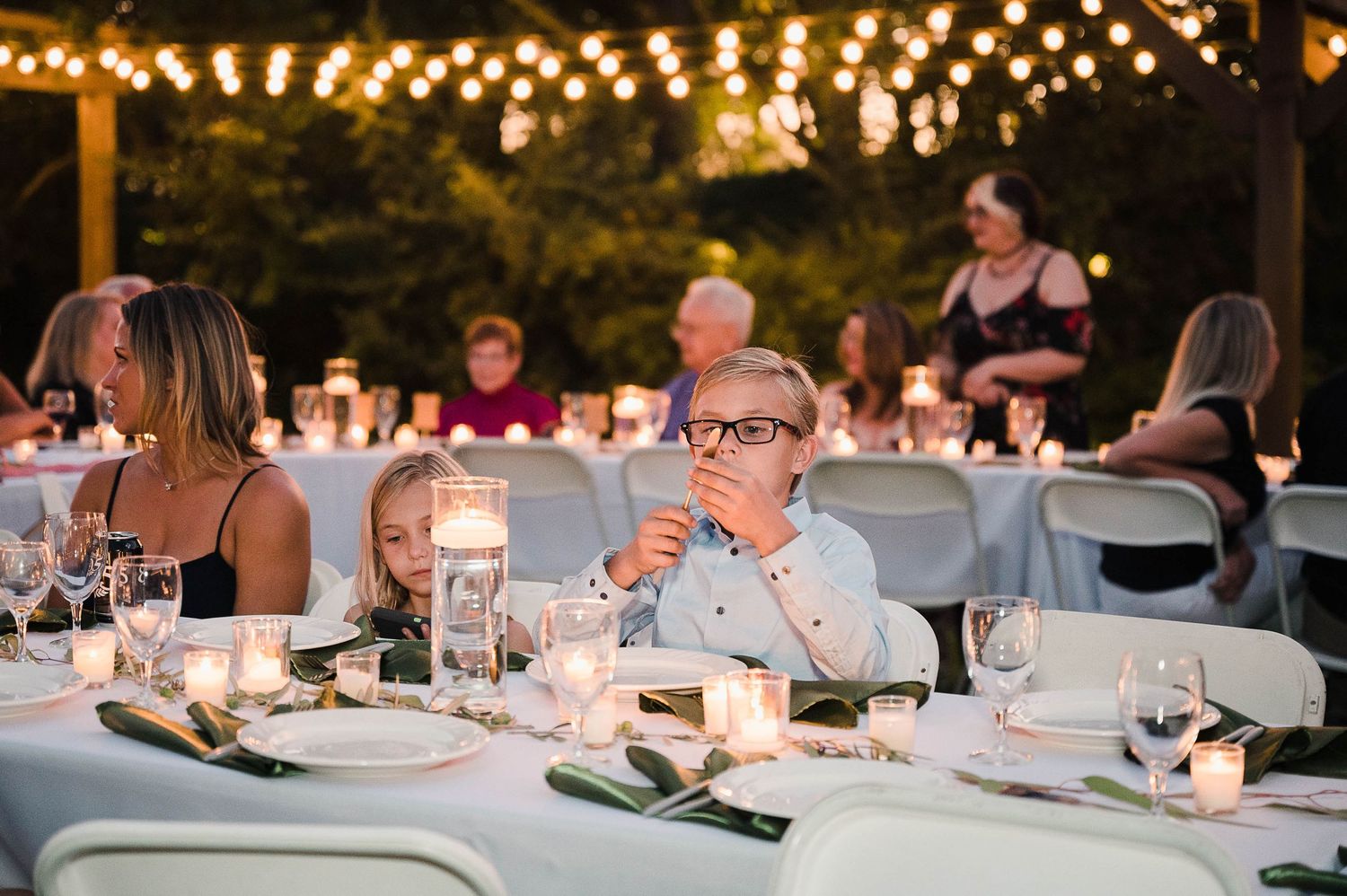 Outdoor reception dining setup with string lights and white tablecloths at dusk.