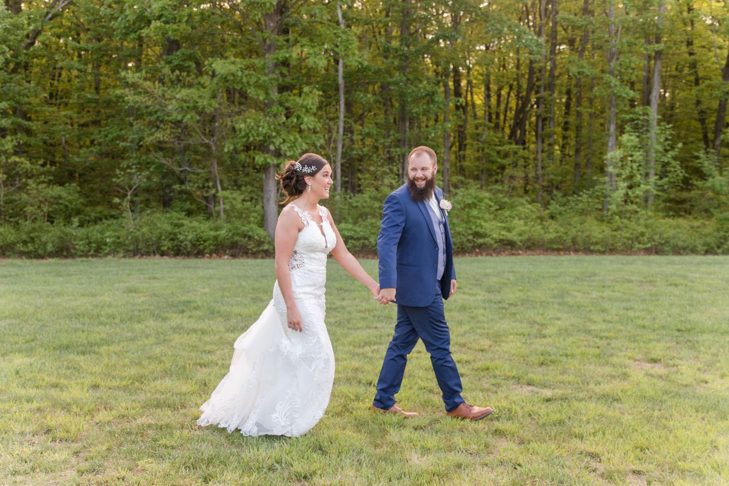 Wedding couple poses playfully on grassy field with forest backdrop.