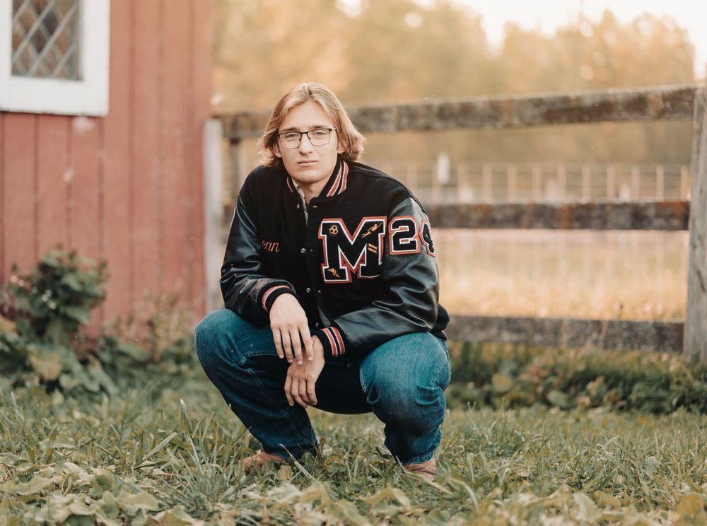Person in varsity jacket crouching casually by red barn in autumn setting.