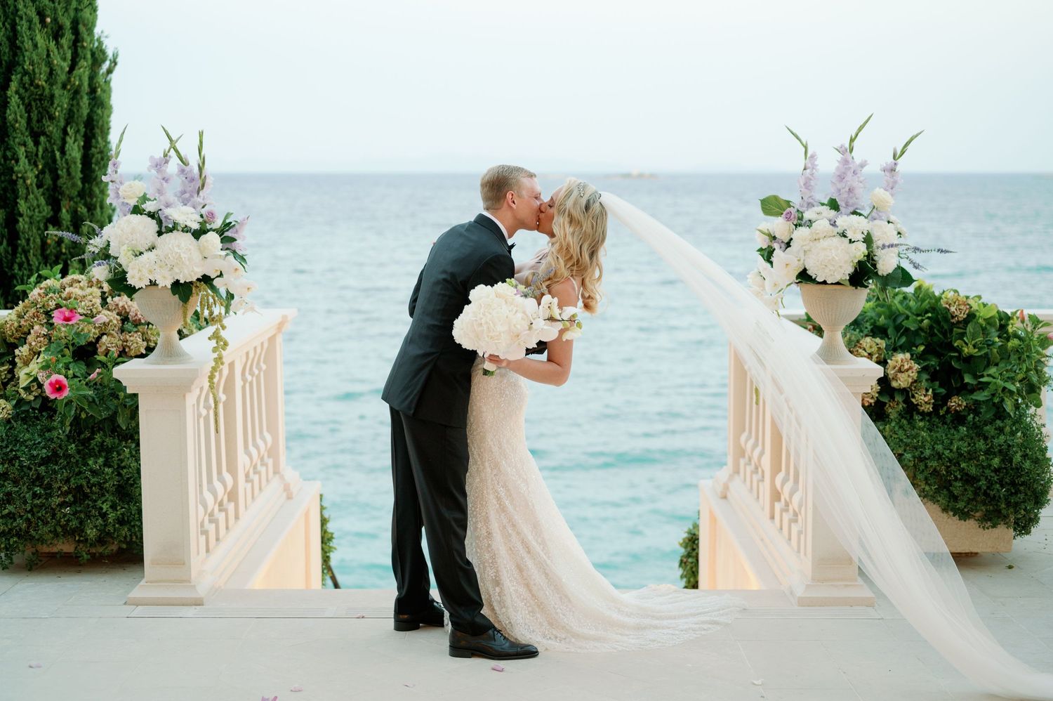 Romantic wedding kiss on oceanfront terrace with floral arch decorations.