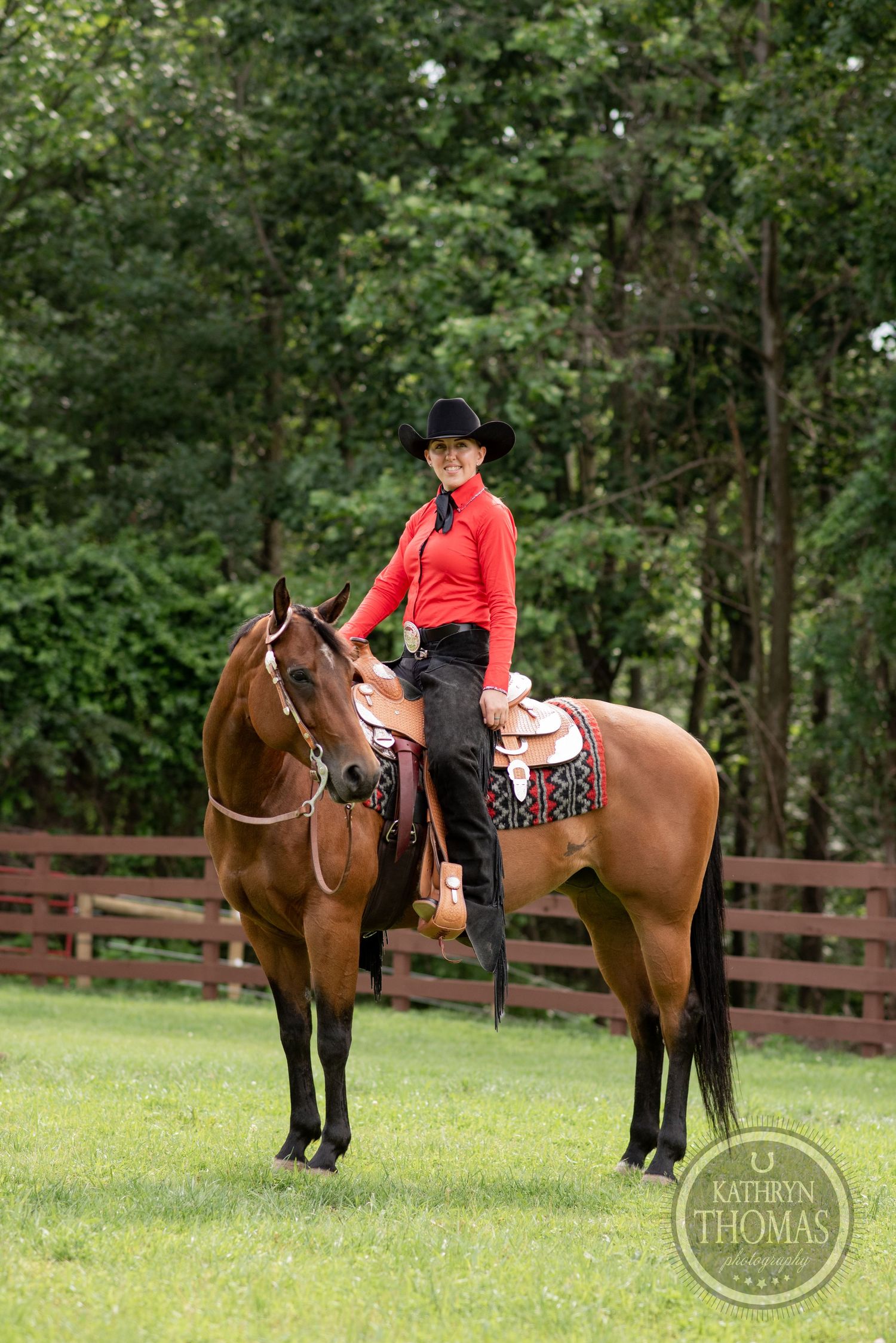 Western rider in red shirt and black hat mounted on chestnut horse by wooden fence.