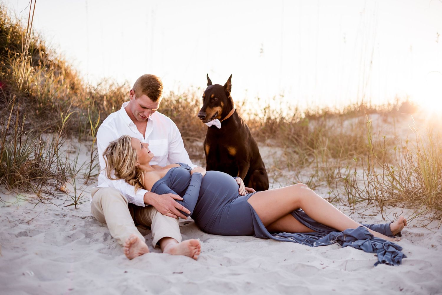 Expecting couple relaxing on beach with brown dog at sunset.
