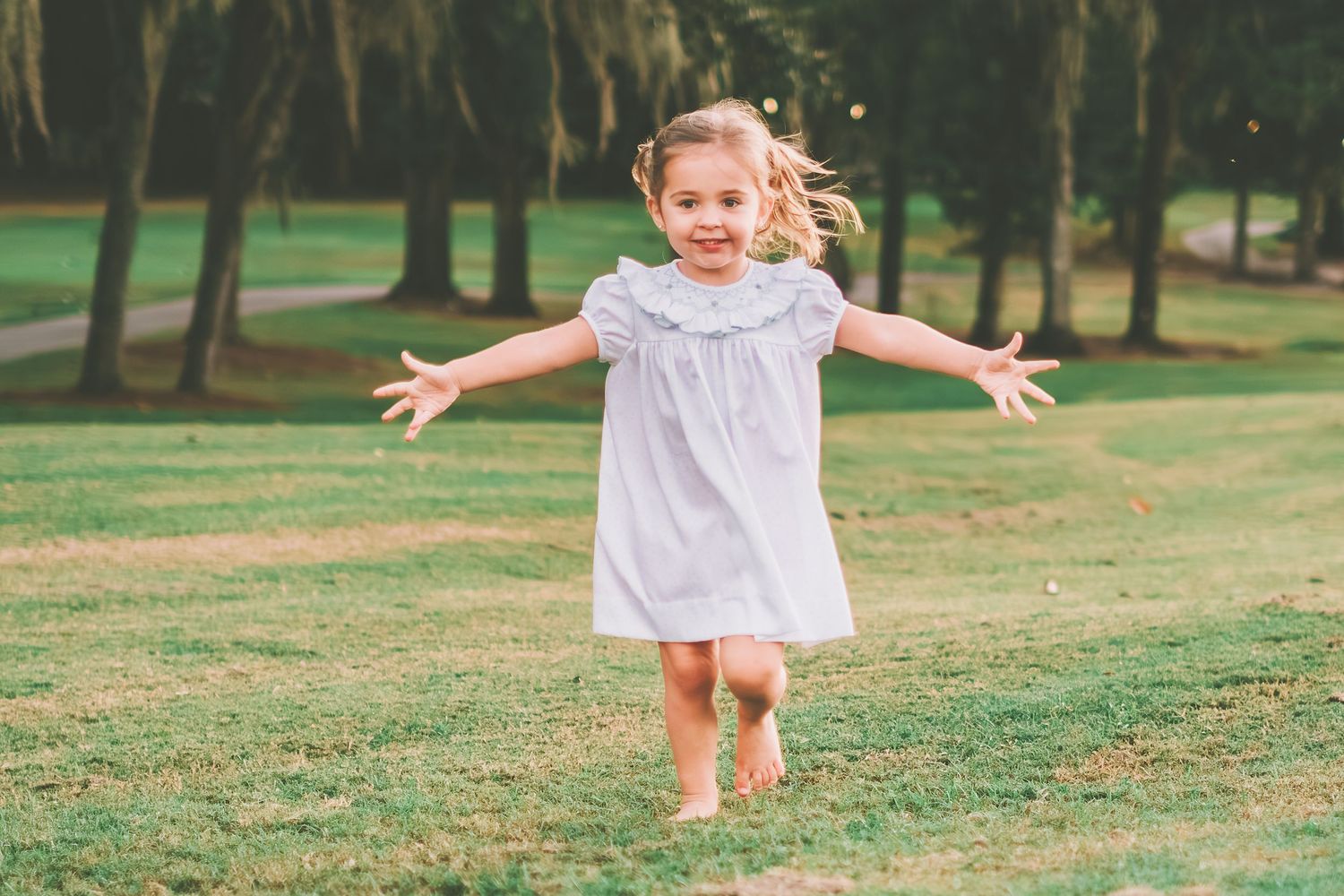 A child runs joyfully through a grassy park wearing a white dress on a sunny afternoon.