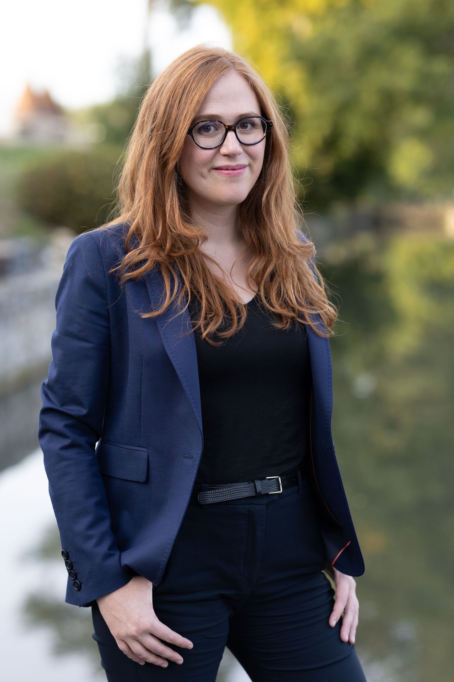 Professional portrait of someone with red hair wearing glasses and a navy business suit standing outdoors by water.