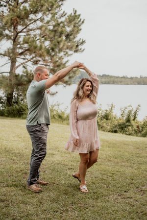 A couple dancing and twirling on grass near a lakeside with pine trees.