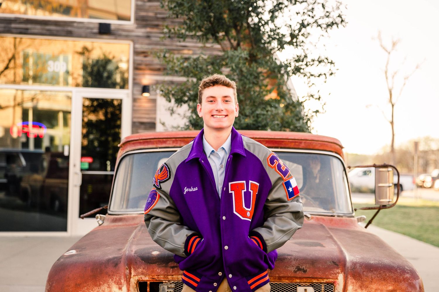 Senior boy in purple varsity jacket poses in front of rustic red truck.