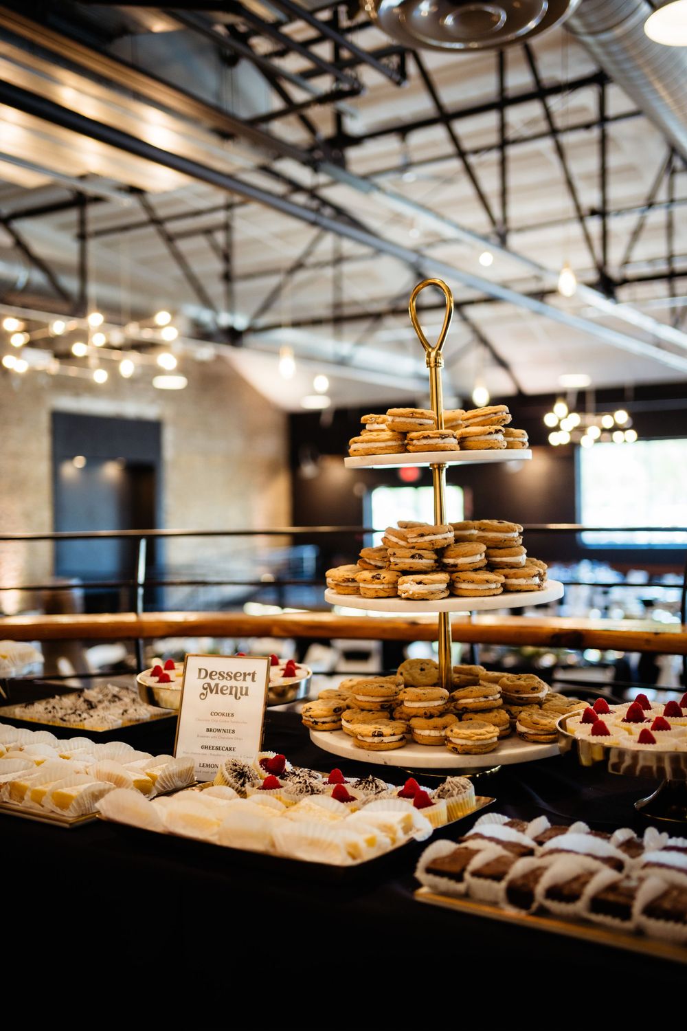 Elegant dessert display featuring tiered stands of pastries and sweets at an upscale event venue with industrial lighting.