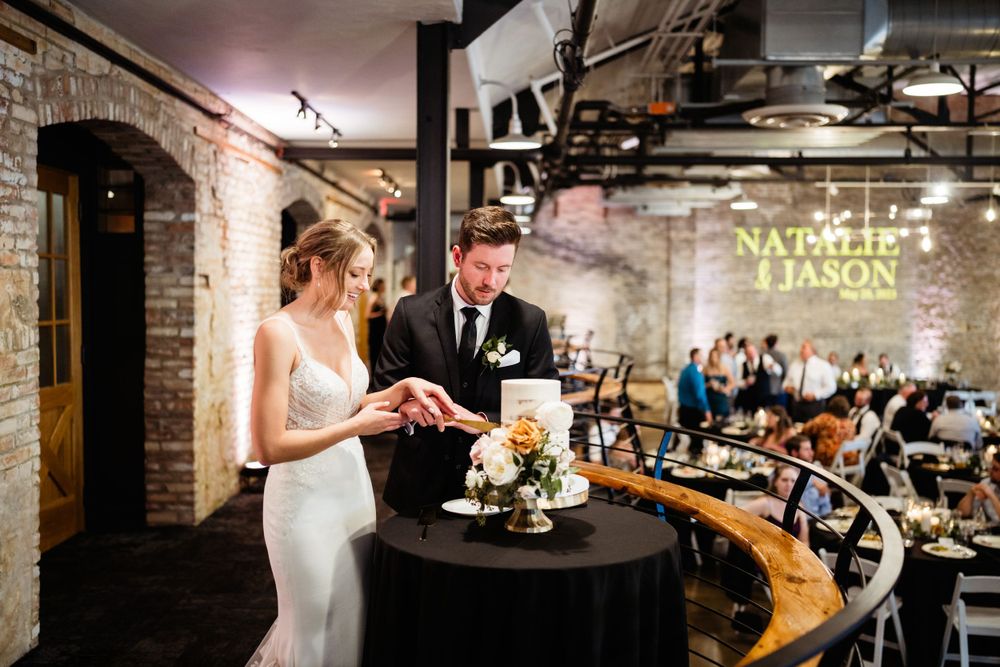 Wedding cake cutting moment at rustic indoor venue with exposed brick walls.