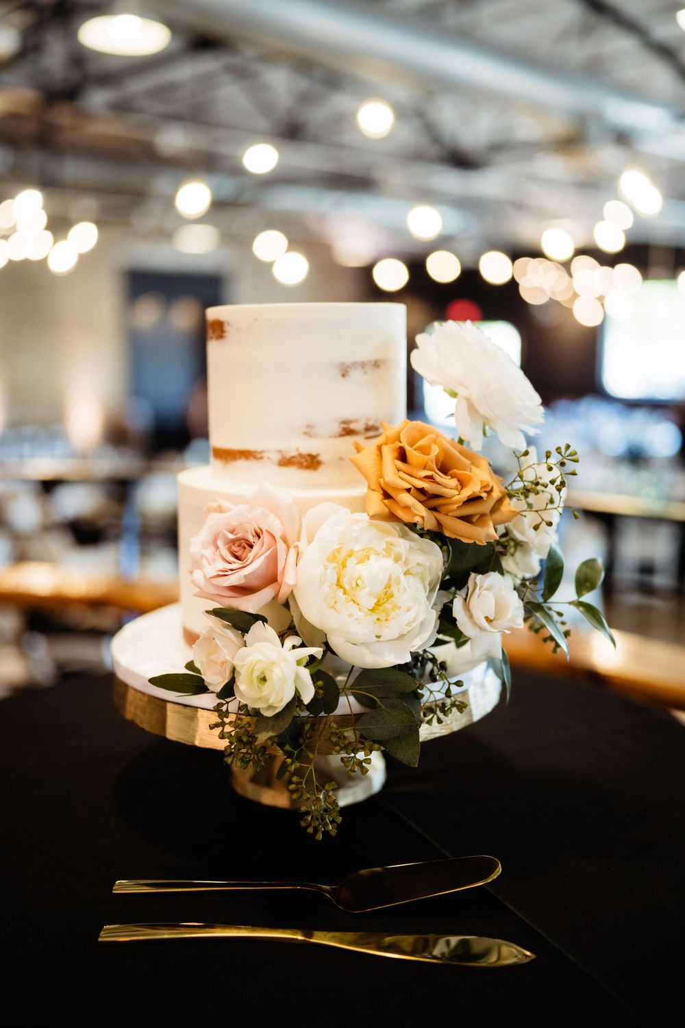 White wedding cake decorated with peach and cream roses on a black tablecloth under industrial lighting.