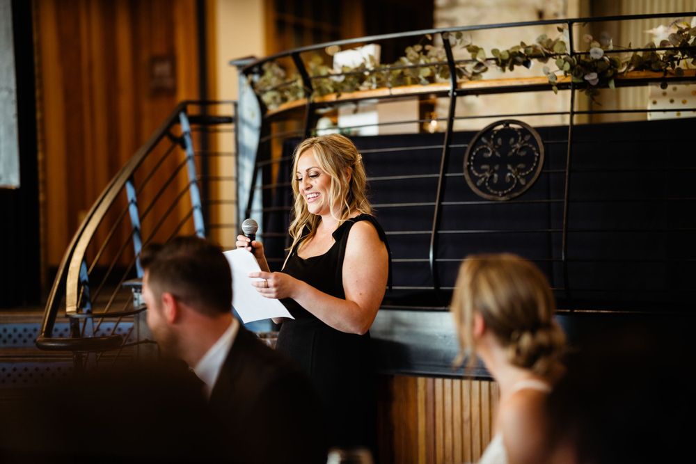 Event speaker delivering speech at venue with decorative railings and wooden interior.