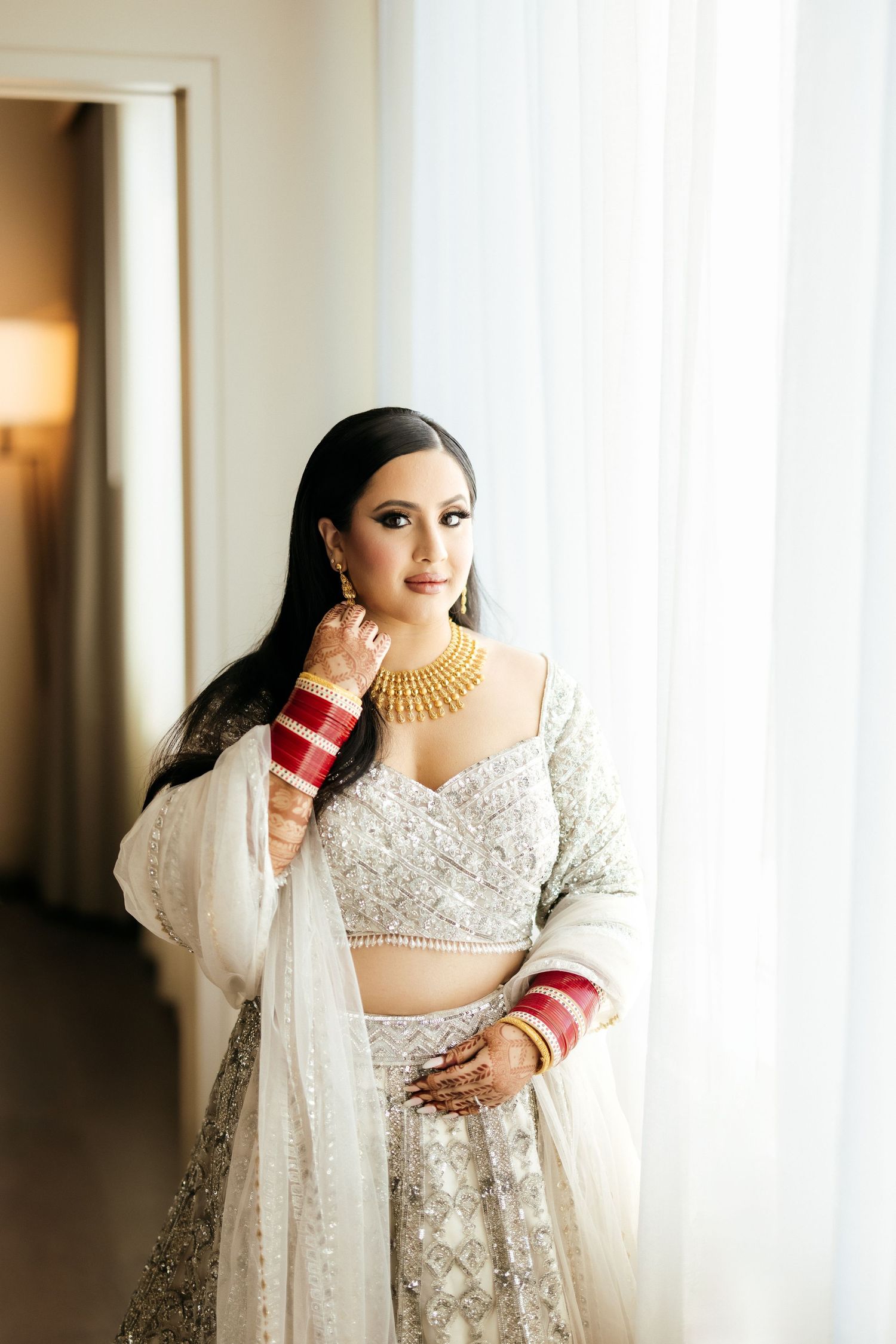 Bride in traditional white and silver South Asian wedding attire standing by window.