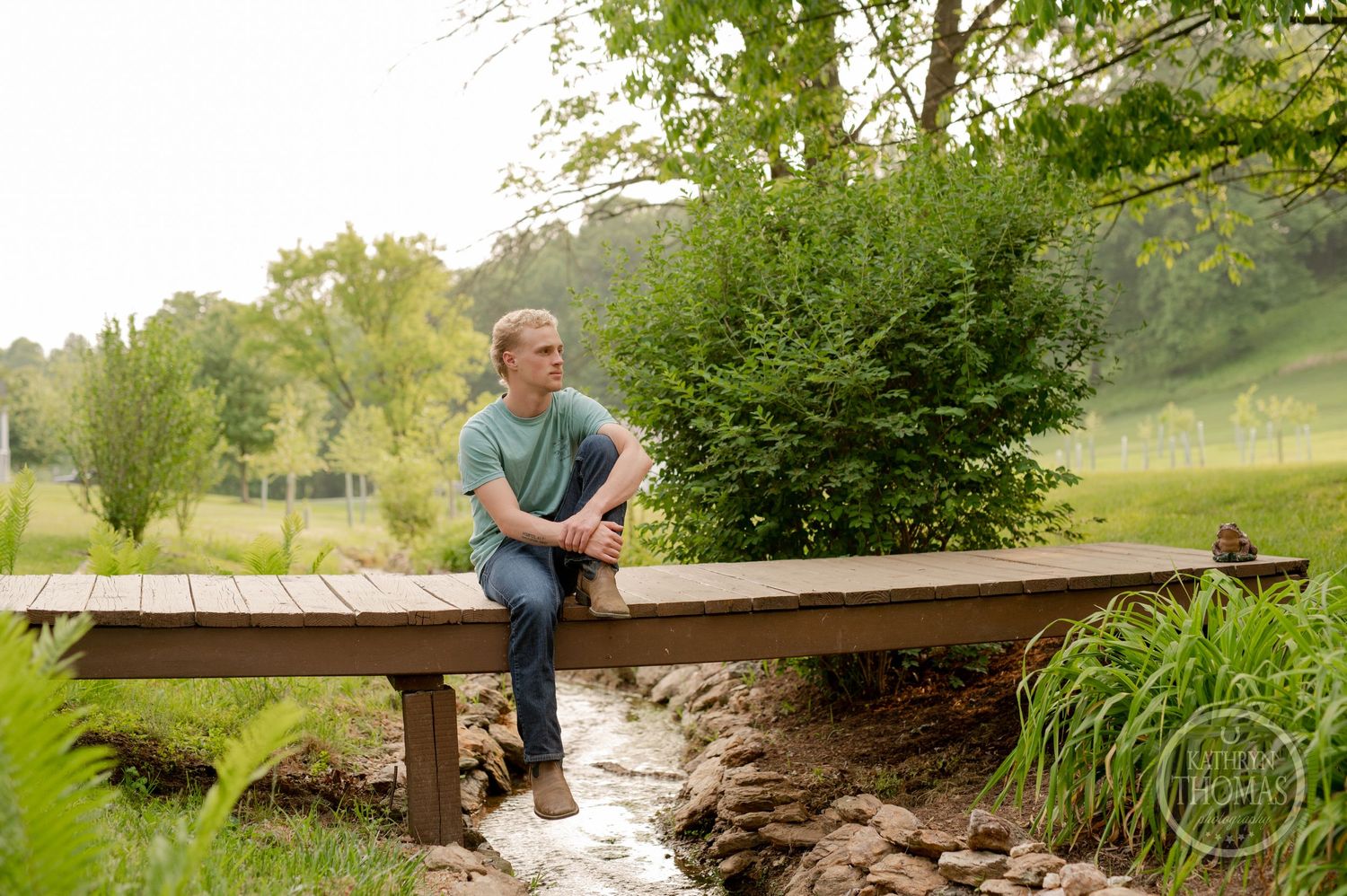 A person sits casually on a wooden bridge over a small stream surrounded by lush greenery.