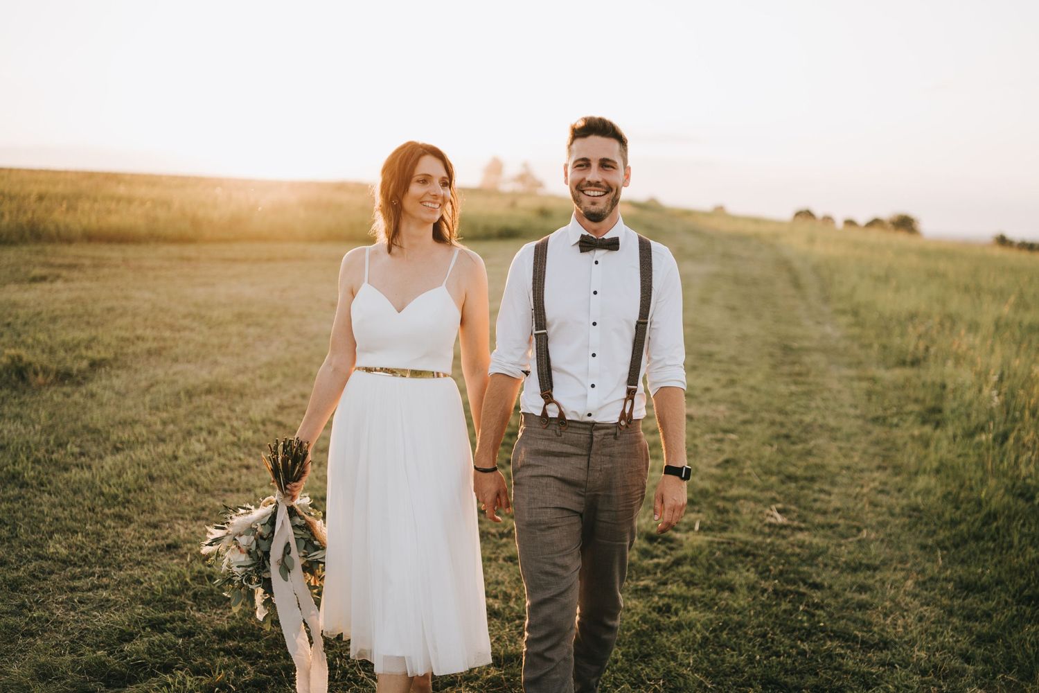Couple walks through a rustic field at sunset in casual wedding attire.
