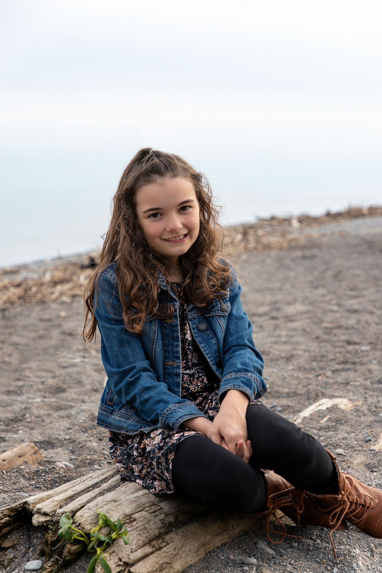 A girl sitting on a log at the beach wearing a jean jacket