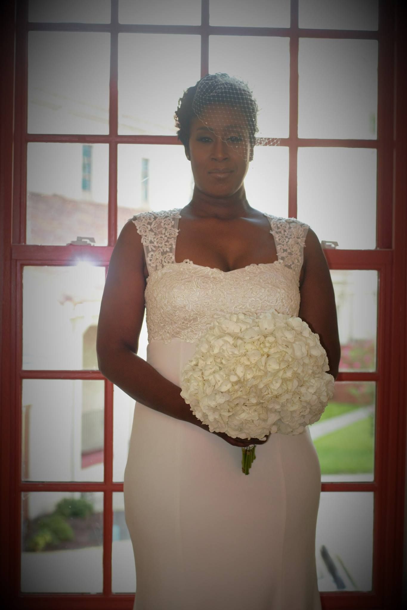 Bride in form-fitting white dress holding large white hydrangea bouquet by window.