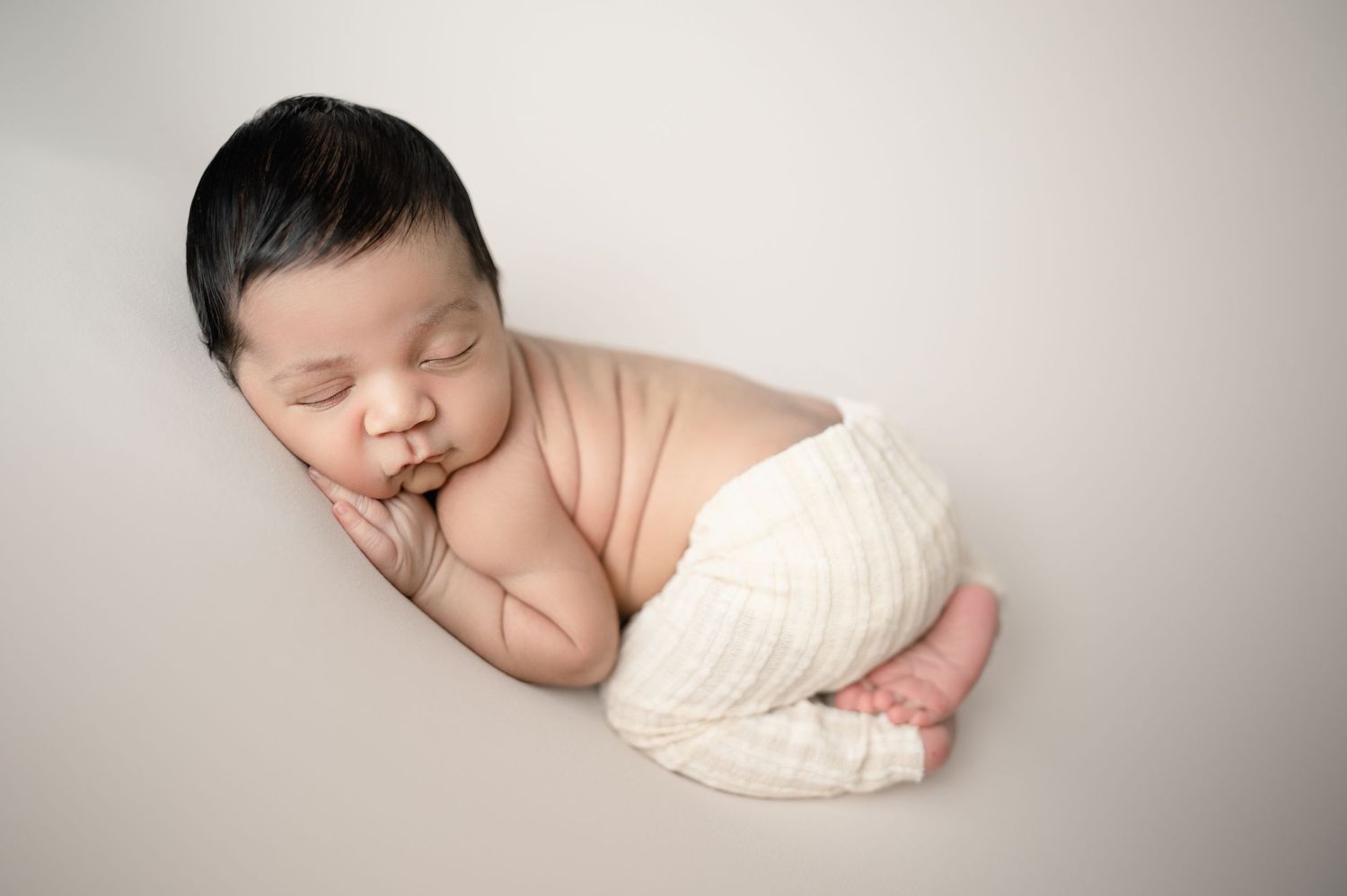 Sleeping newborn in white diaper resting on cream backdrop.