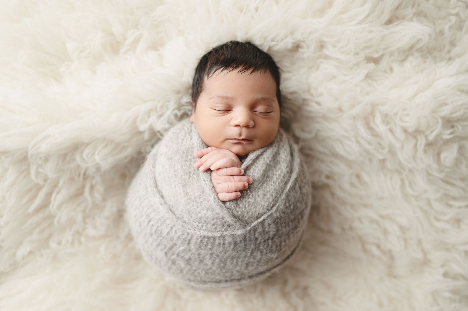 Newborn wrapped in gray knit fabric lying on plush white fur background.