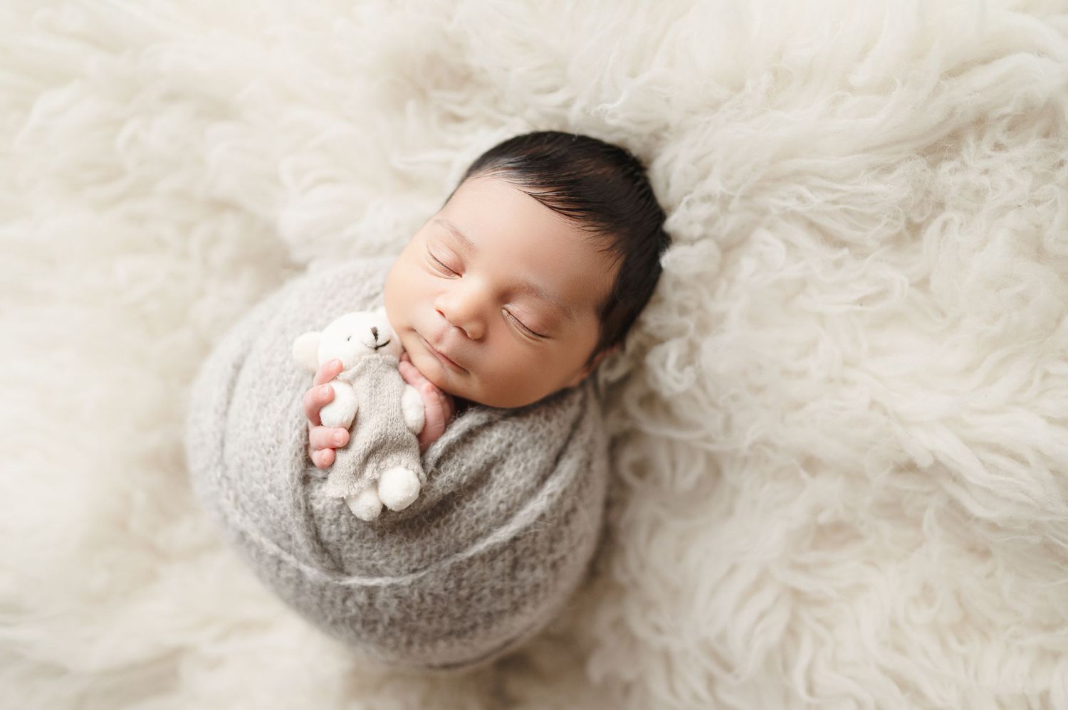 Peaceful newborn photography with soft gray wrapping on white furry backdrop.