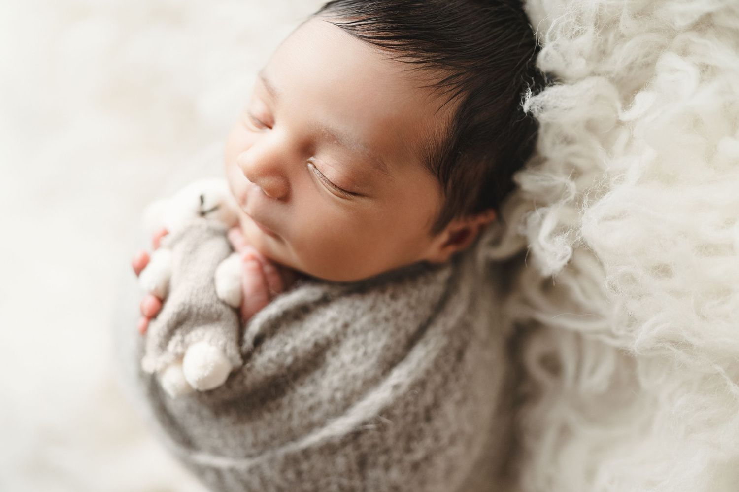 Sleeping newborn nestled in gray knit wrap on cream textured background.