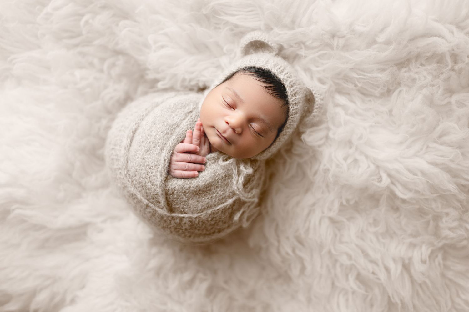 Newborn baby wrapped in beige knit blanket lying on white textured backdrop in professional photo studio setting.