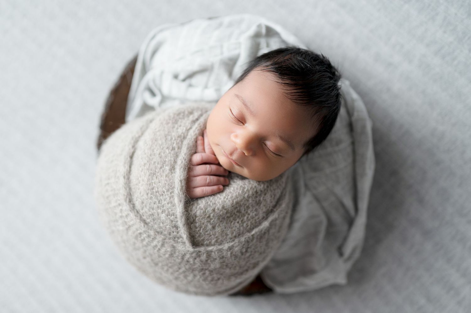 Minimalist newborn photography featuring baby wrapped in light gray blanket on soft white background.