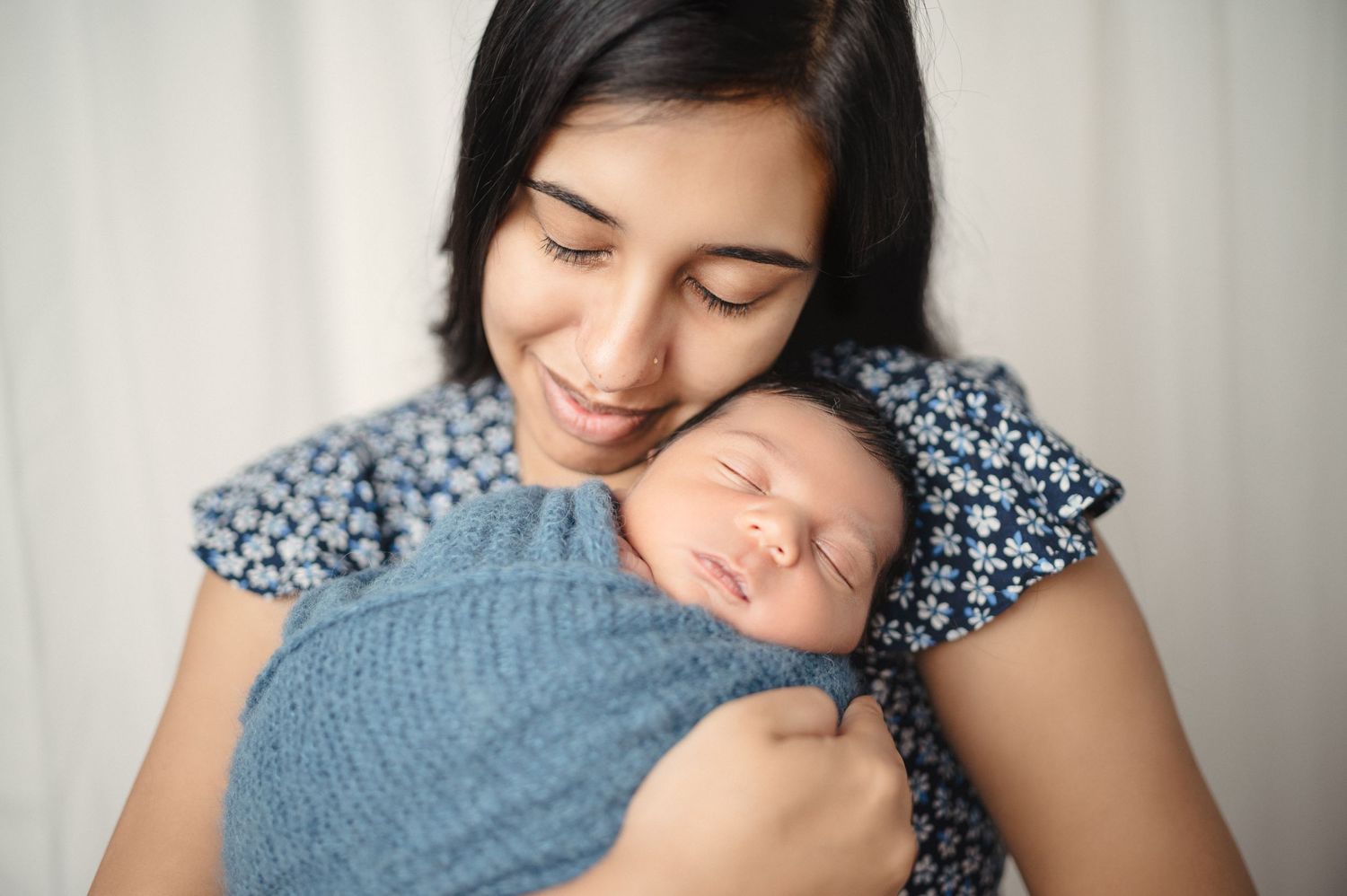 Gentle portrait series showing mother cuddling newborn in blue swaddle against light backdrop.