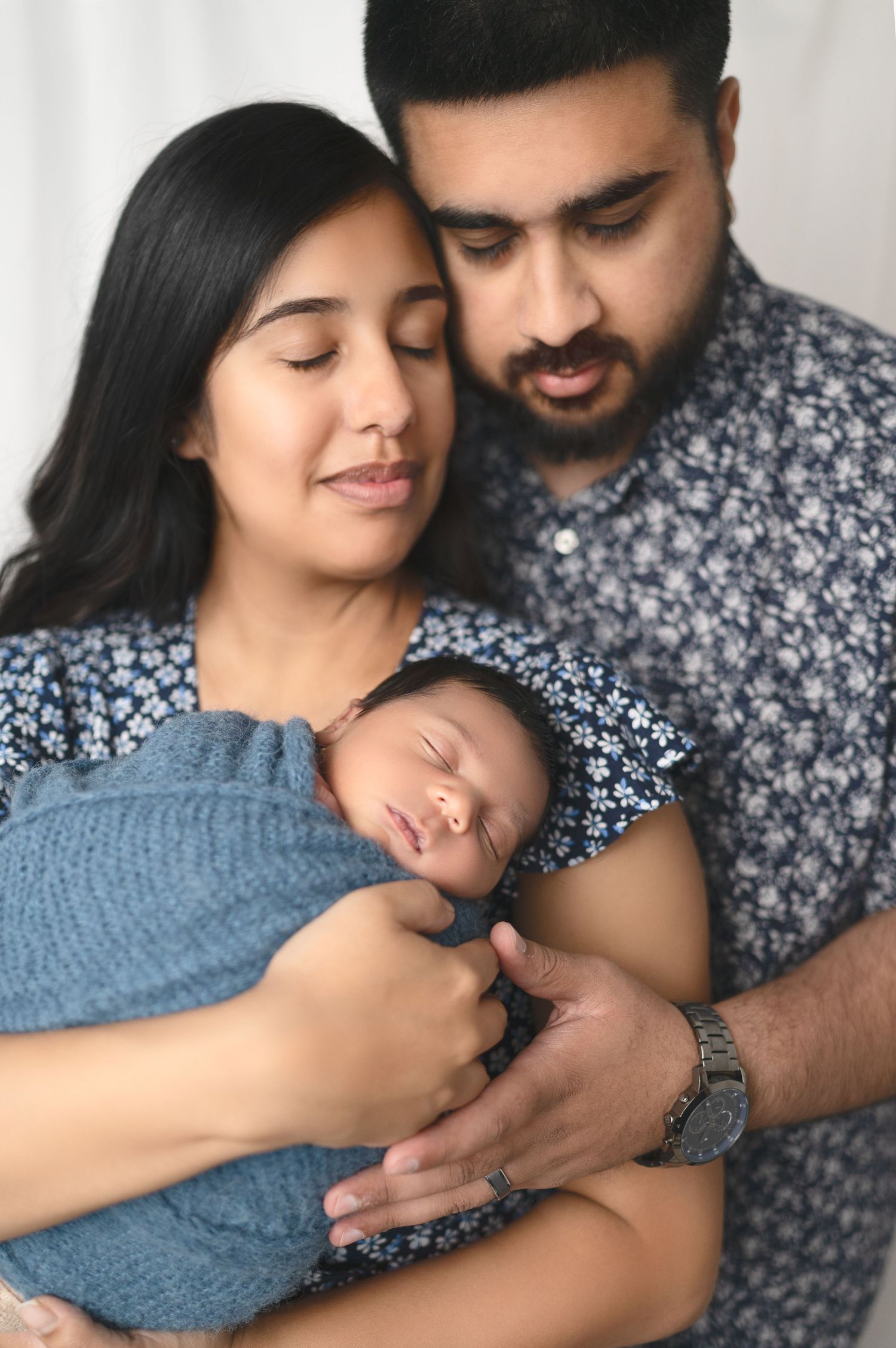 A couple gently embraces their newborn baby wrapped in a blue blanket against a light background.