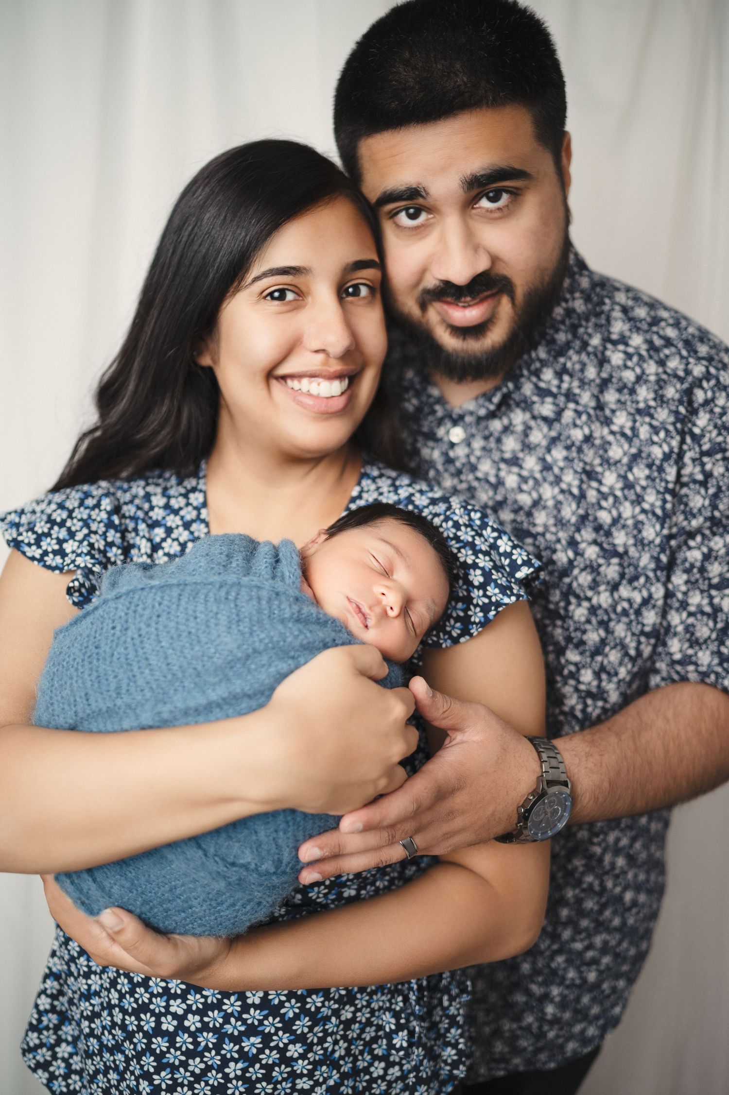 New parents smile while holding their sleeping newborn baby swaddled in soft blue fabric.