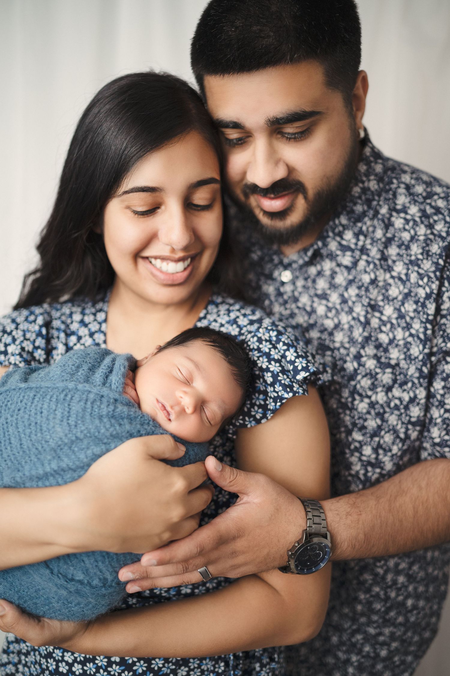 Parents share an intimate moment with their sleeping newborn wrapped in a cozy blue blanket.