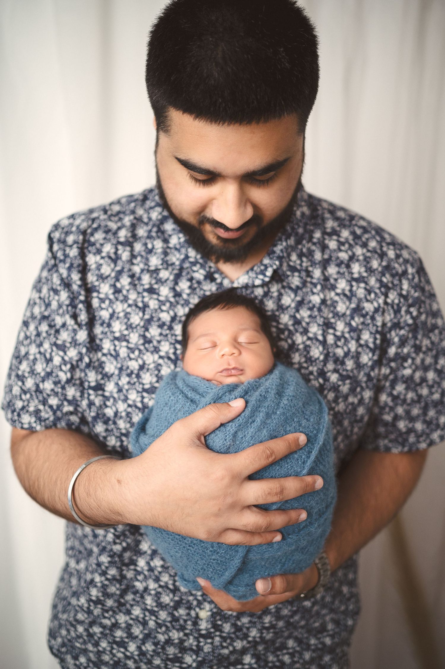 A parent in a floral shirt tenderly holds their sleeping newborn swaddled in blue.
