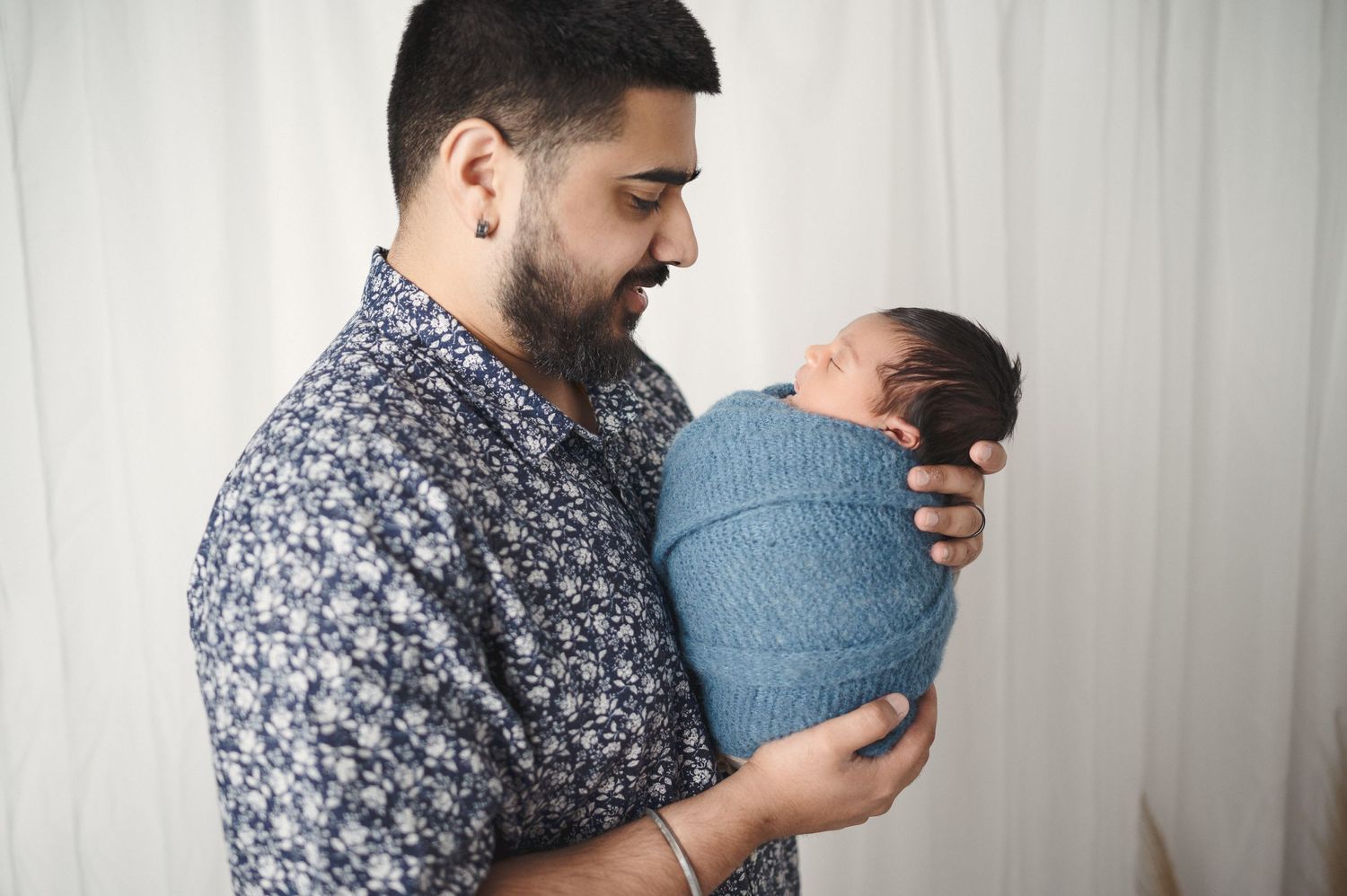 A parent in a floral print shirt gazes lovingly at their newborn swaddled in blue.