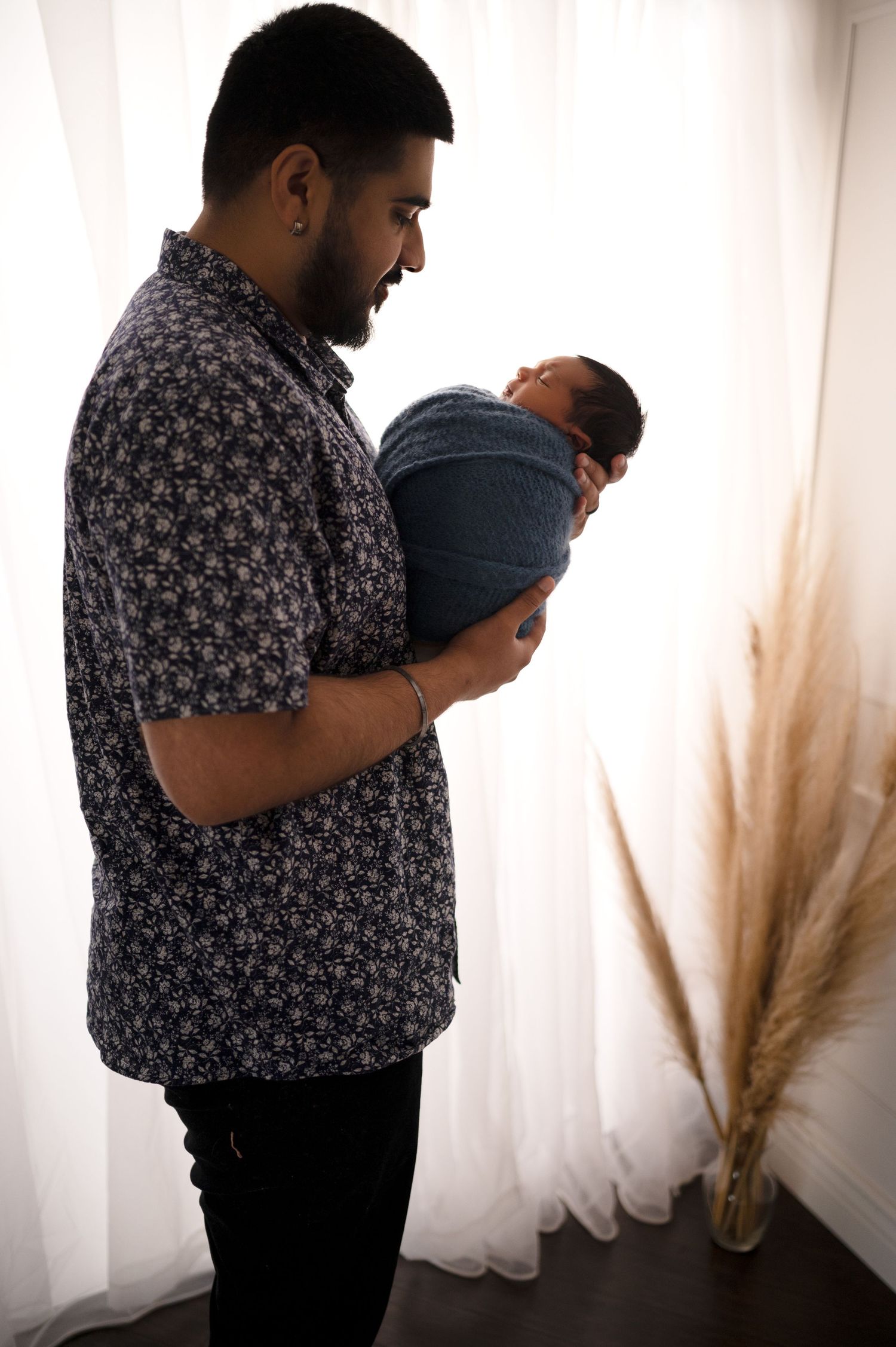 A parent holds their sleeping newborn beside white curtains and pampas grass decor.