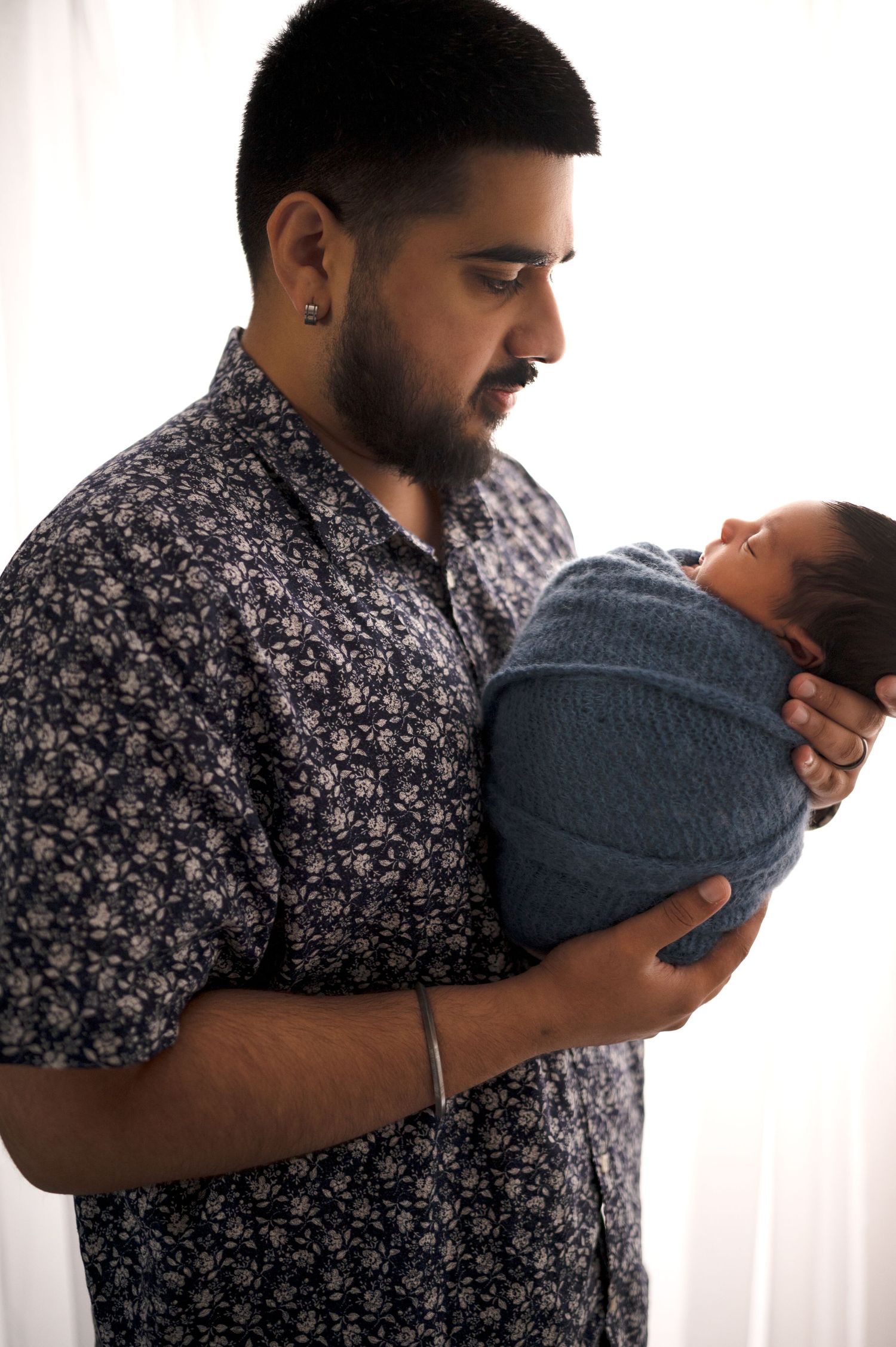 A parent in a floral shirt looks down adoringly at their swaddled newborn baby.