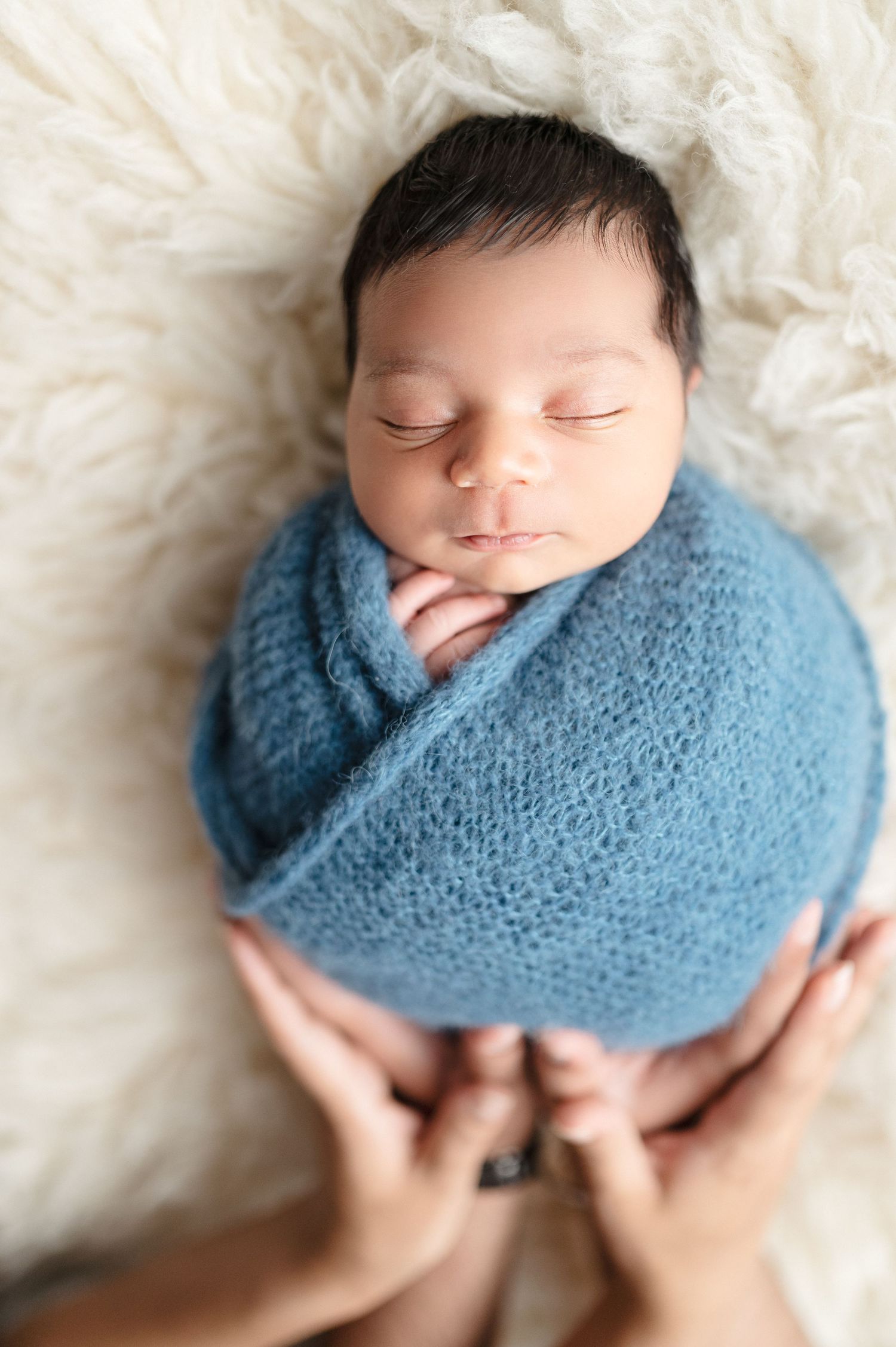 Sleeping newborn wrapped in blue knit blanket rests on cream textured background.