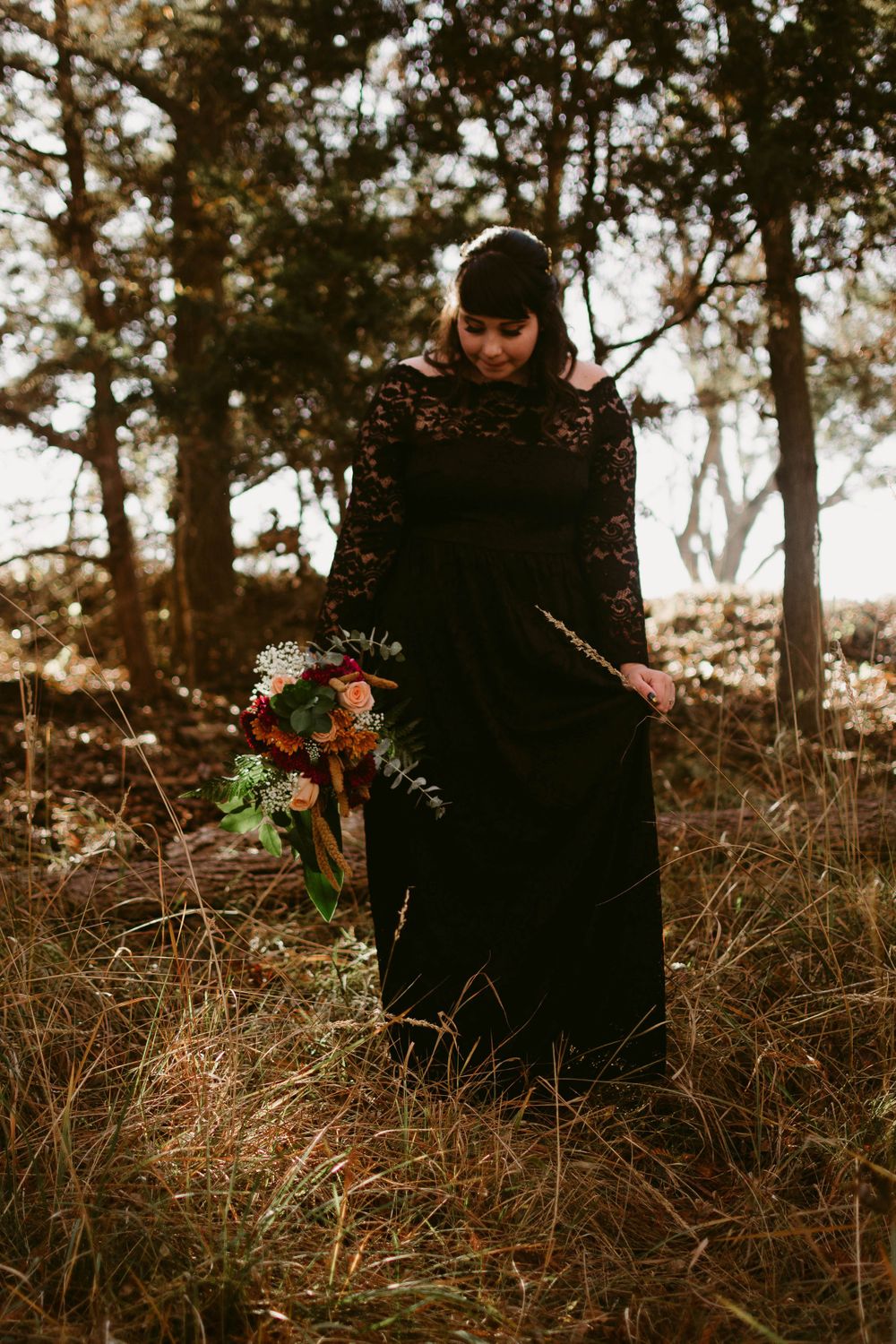 A figure in a black lace dress holds a vibrant bouquet while standing in autumn woods.