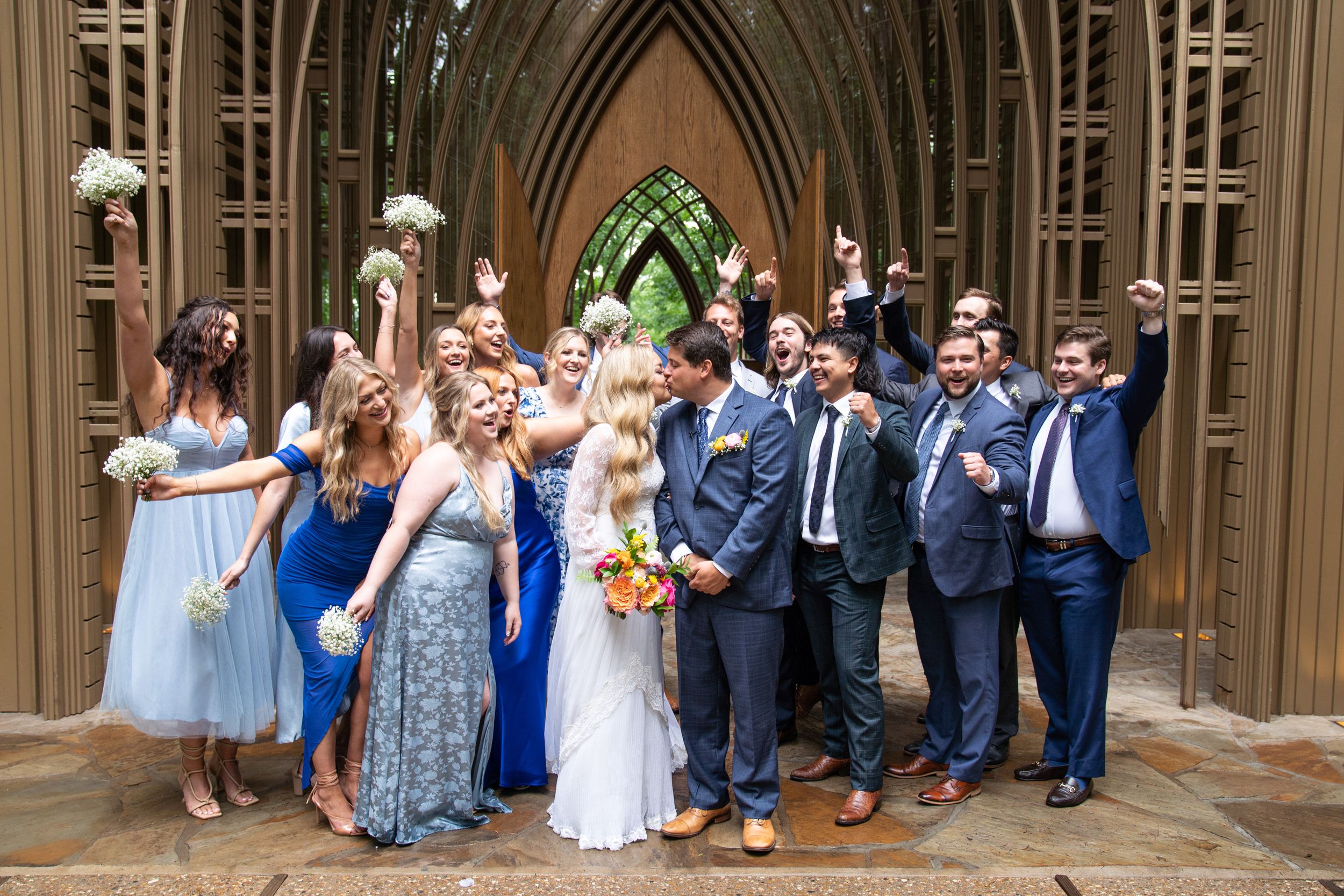 Wedding party group photo under wooden gothic arches outdoors.