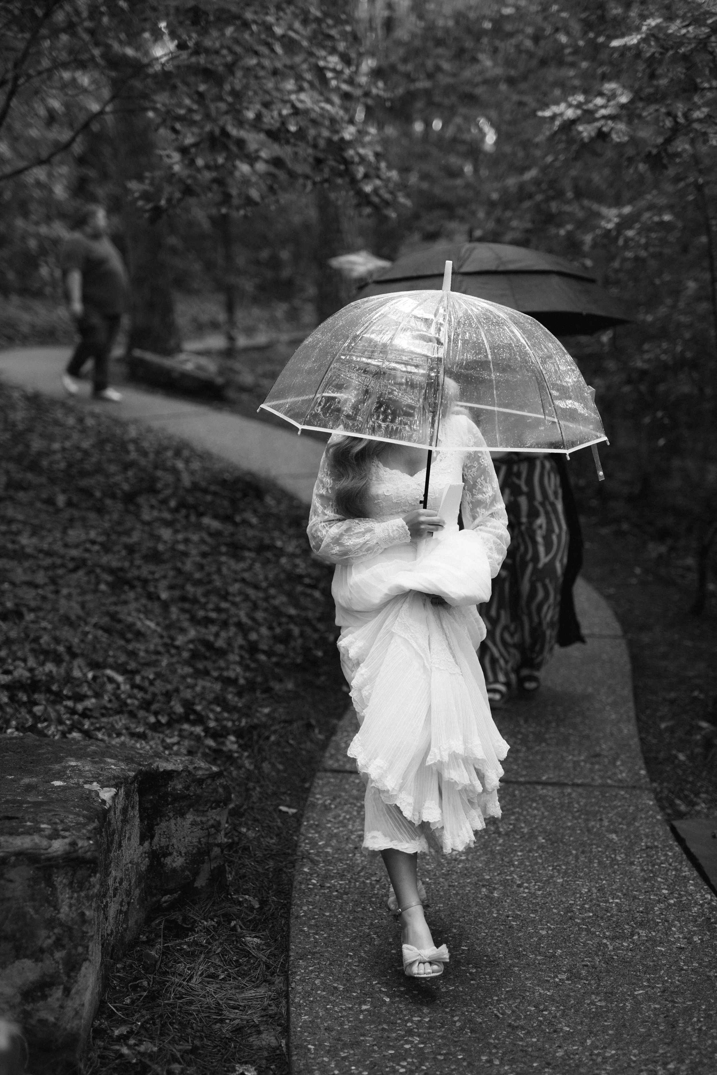 Clear umbrellas shelter figures in white formal attire walking along a rainy garden path.