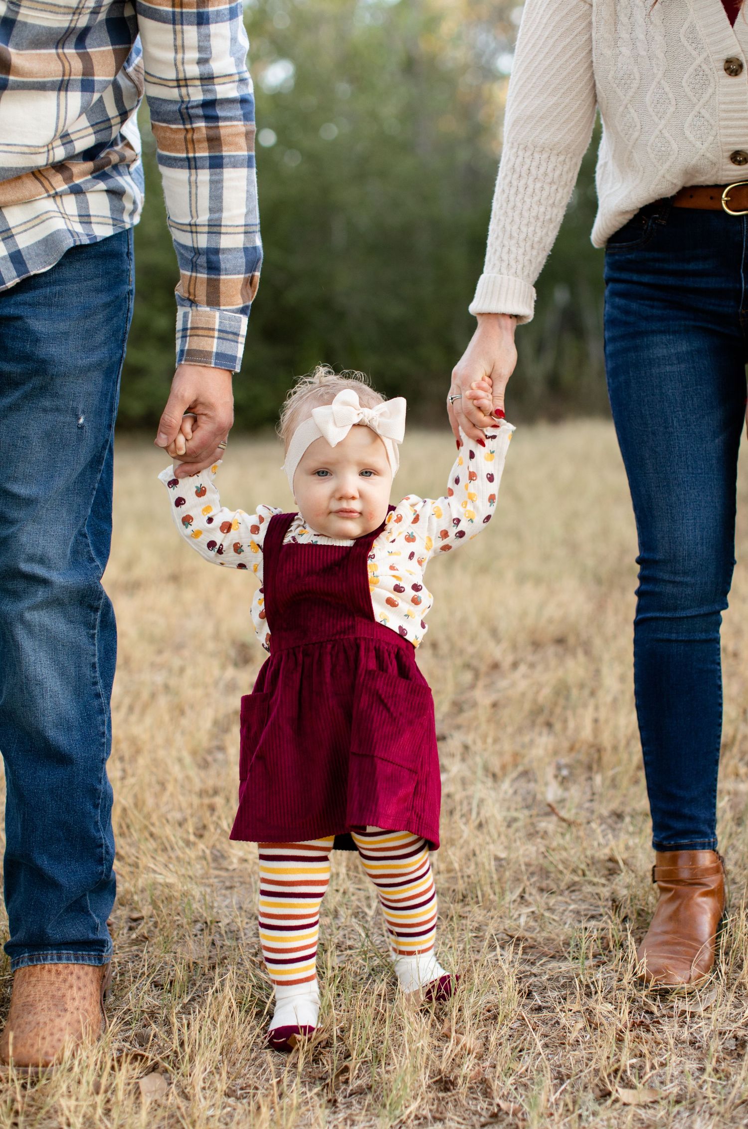 Little one in burgundy jumper dress and striped tights walking while holding parents' hands.