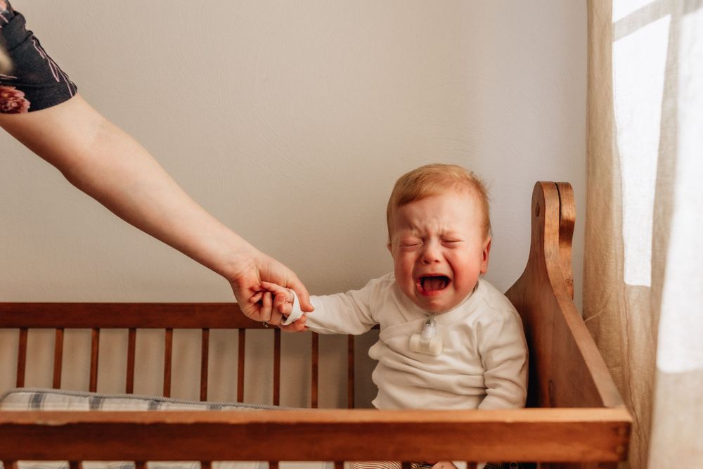 toddler crying in crib with mothers hand coming in from the left hand side of the frame