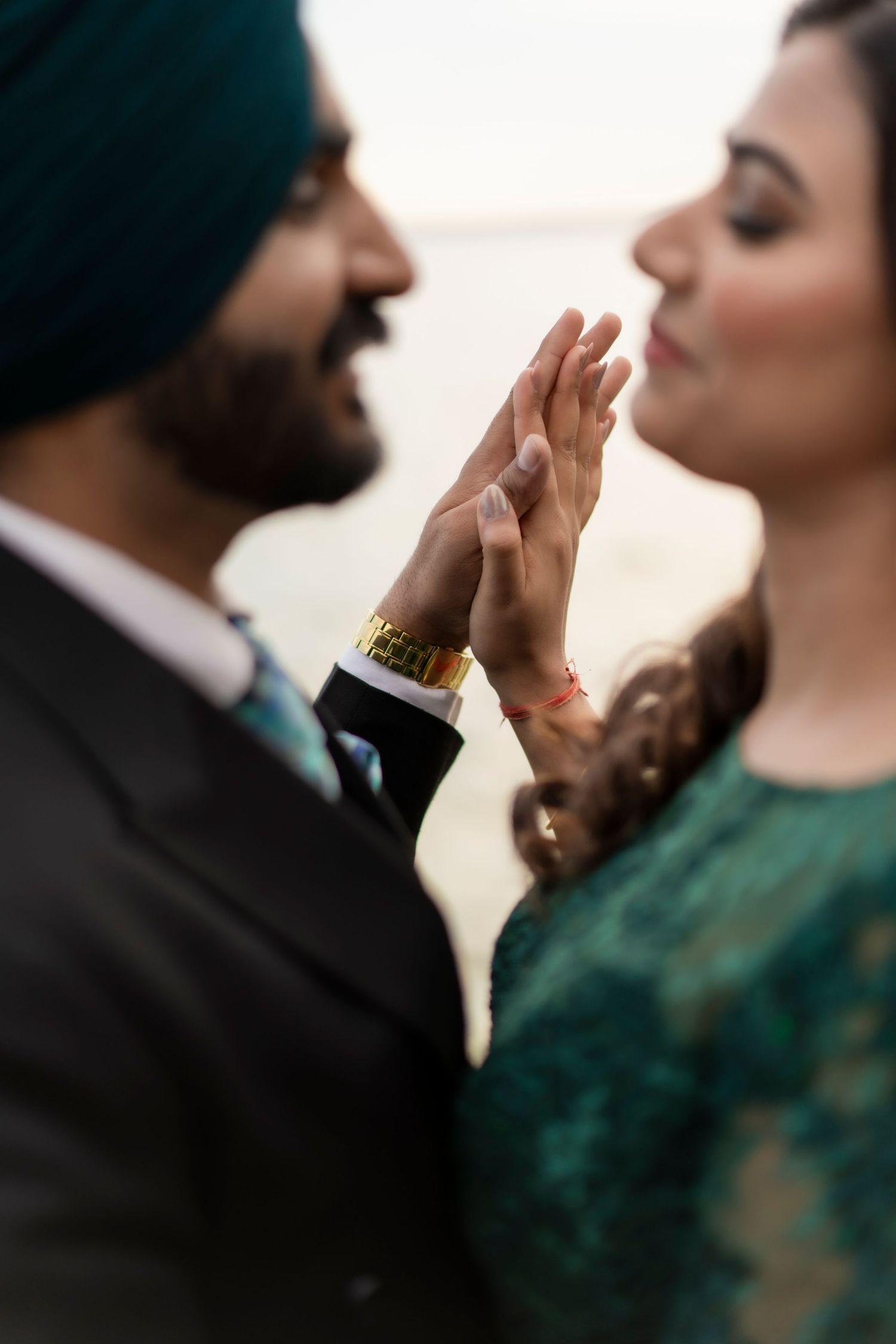 A couple in formal attire share an intimate moment with their hands touching, with a dark turban and green dress visible.