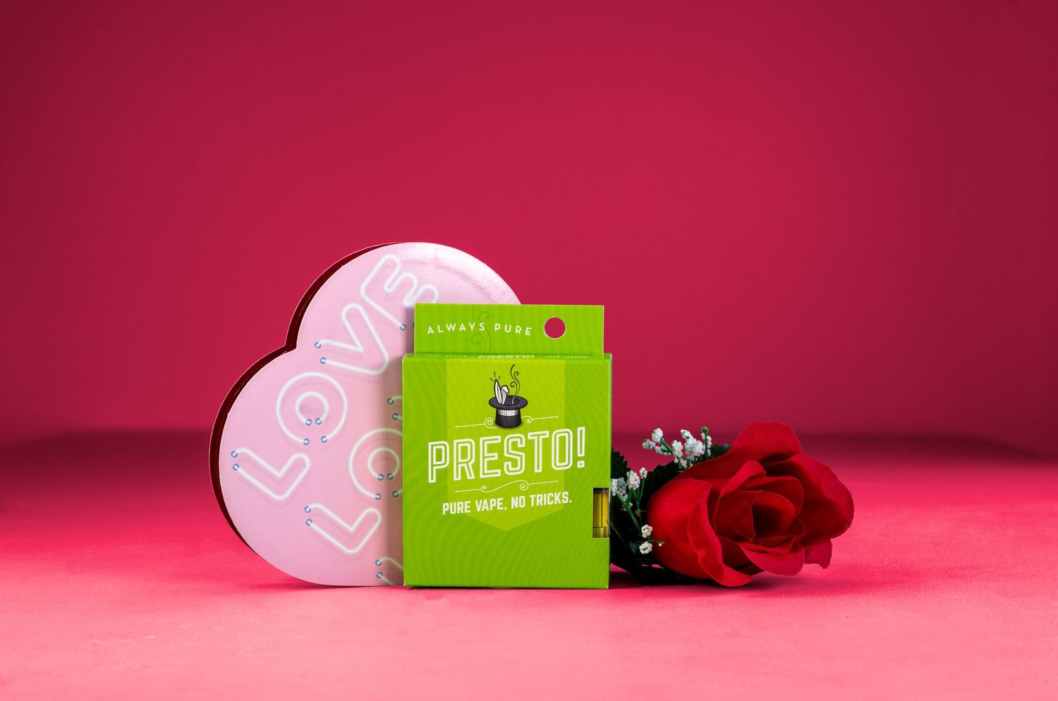 a product box sits on a pink background in front of a box of chocolate and a rose.