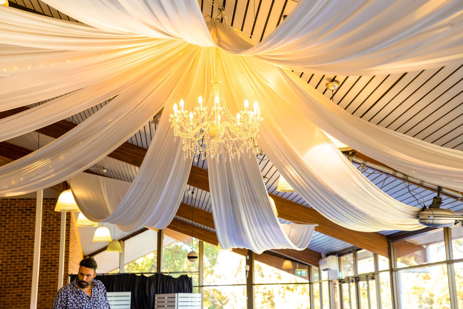 Wedding reception space with elegant white fabric ceiling draping and crystal chandeliers creating soft lighting.